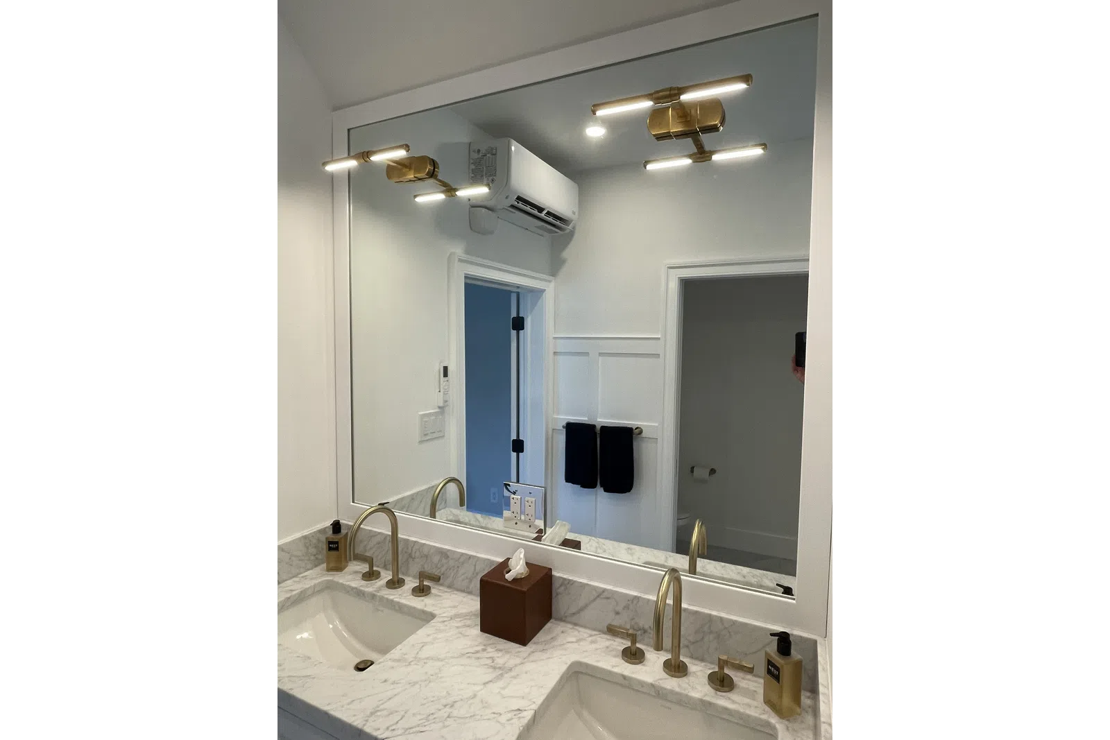Modern bathroom with dual sinks, marble countertop, brass faucets, large mirror, and wall-mounted air conditioner.