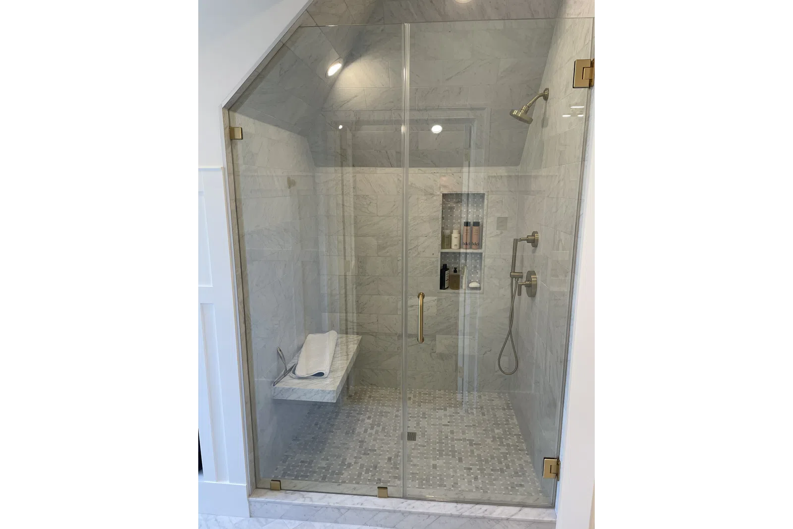 Modern marble tiled walk-in shower with glass doors, built-in bench, and gold-tone fixtures and showerhead.