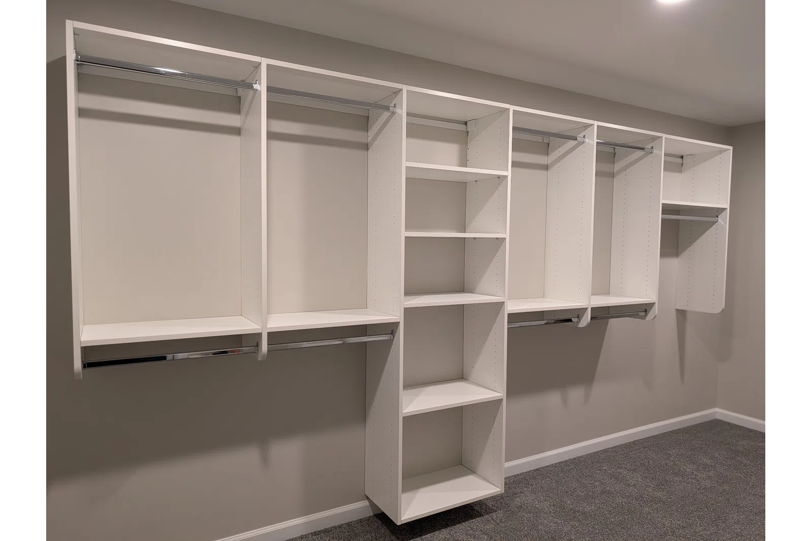 Empty white modular closet organizer with shelves and hanging rods against beige wall and carpeted floor.
