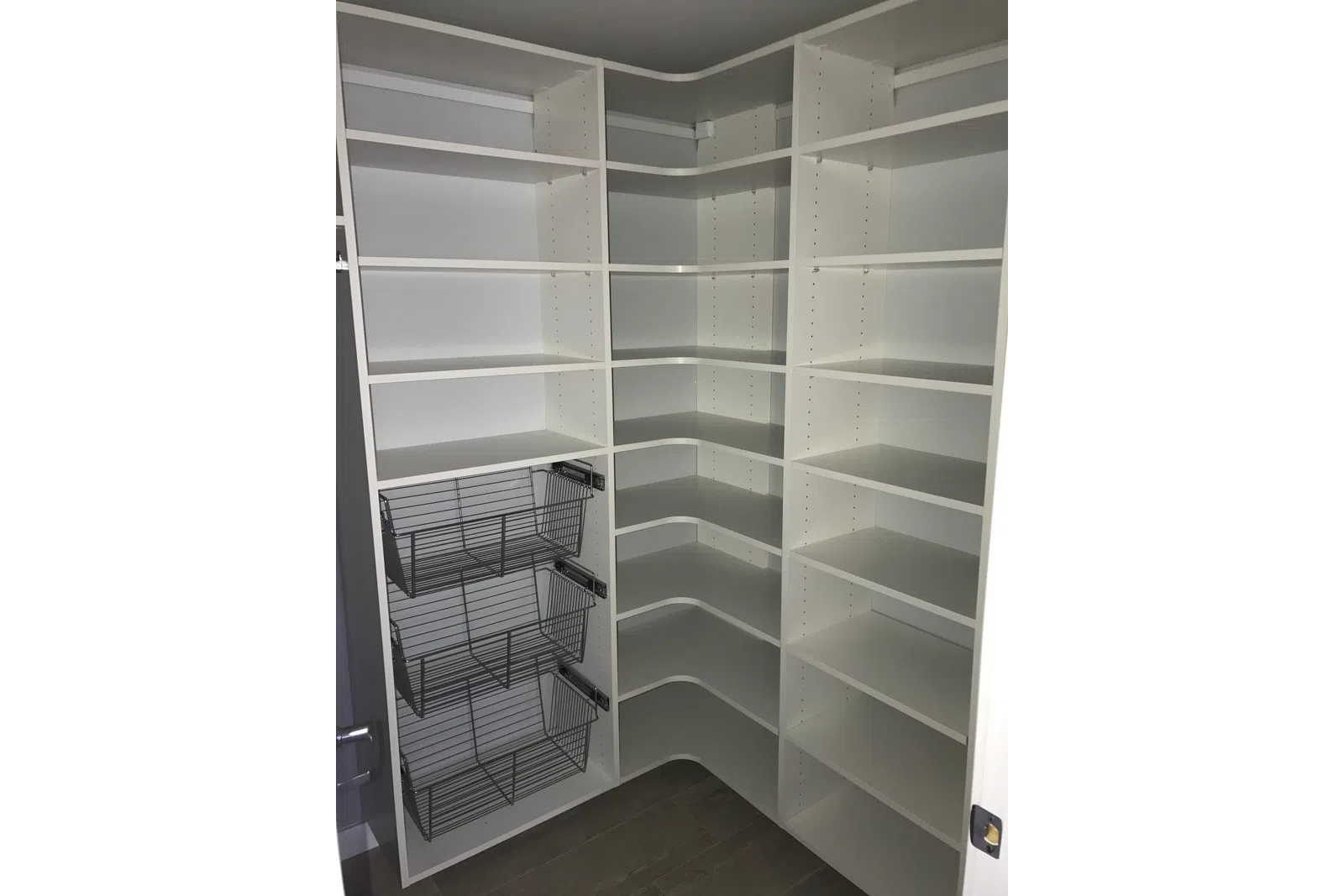 Empty white corner pantry with adjustable shelves and three metal wire baskets on the left side.