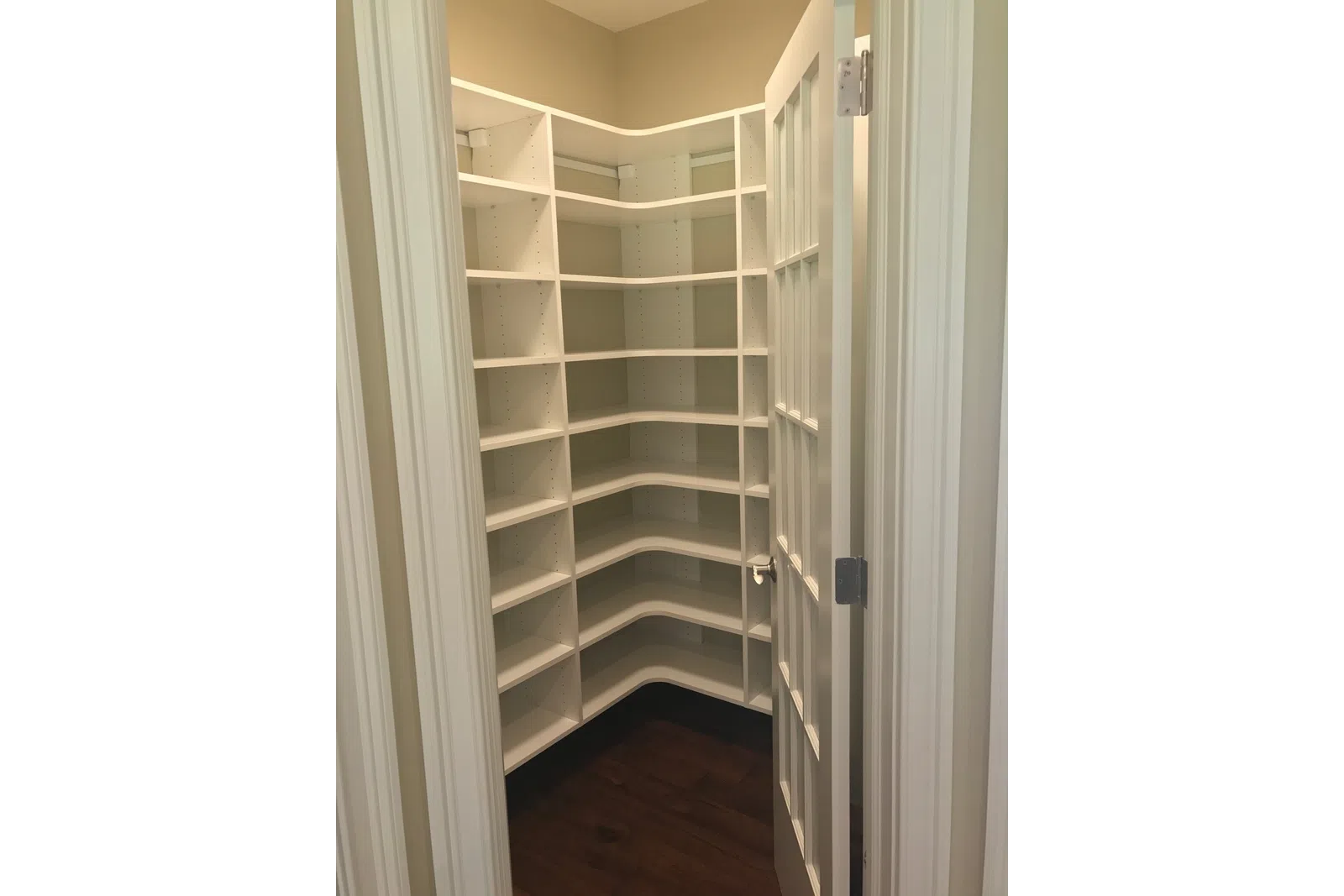 Empty white corner pantry shelves inside a small room with an open door and dark wooden floor