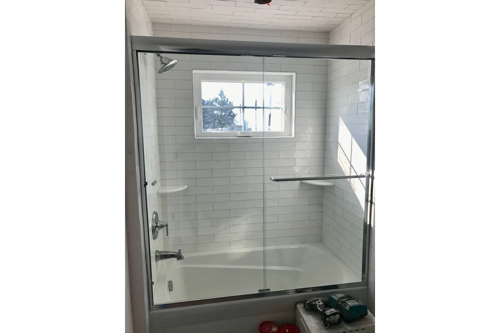 Clean modern white tiled bathtub with glass shower door and a window letting in natural light.
