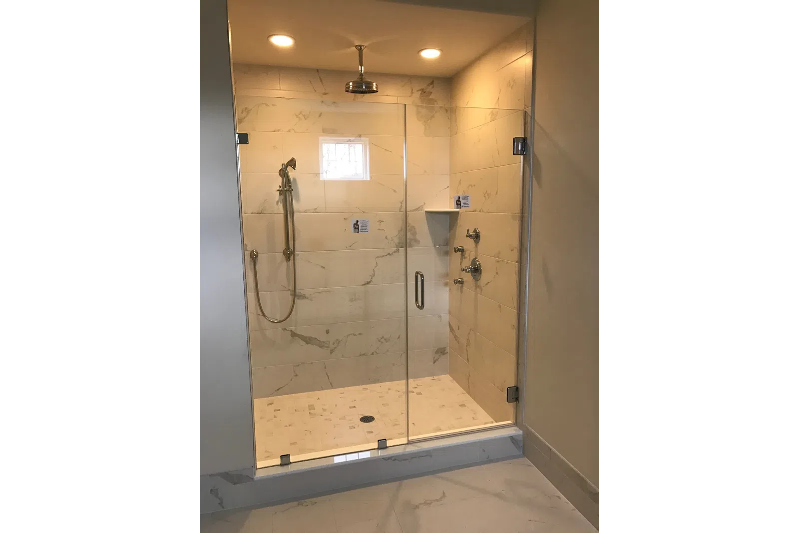 Modern walk-in shower with glass door, marble tiles, rain showerhead, and handheld shower in bathroom.