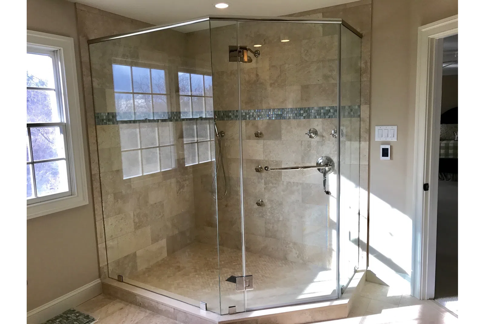 Modern frameless glass shower enclosure with beige tile walls and blue mosaic accent strip in bright bathroom.