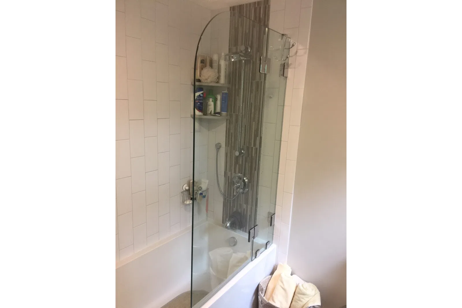Modern bathroom with glass shower screen, bathtub, tiled wall, showerhead, and toiletries on corner shelves.