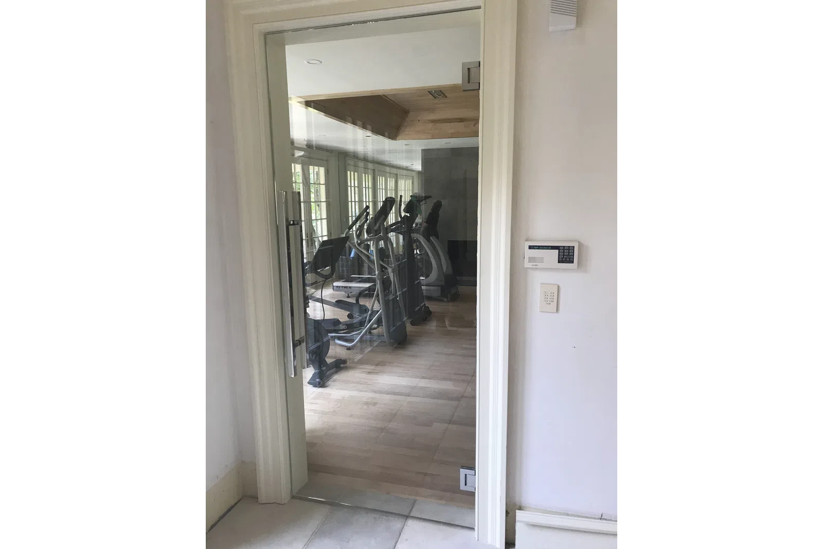 View of a home gym with exercise equipment behind a glass door and security keypad on wall.