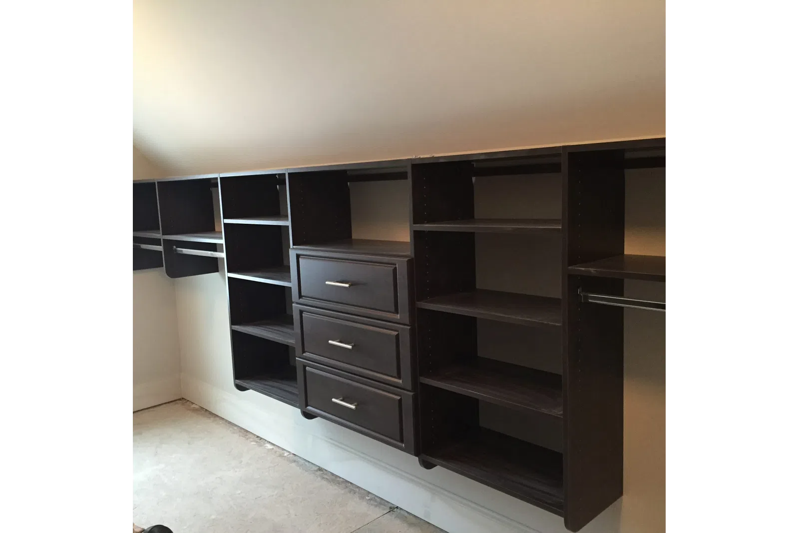 Dark wood floating closet shelves and drawers with metal handles under sloped ceiling in empty room.