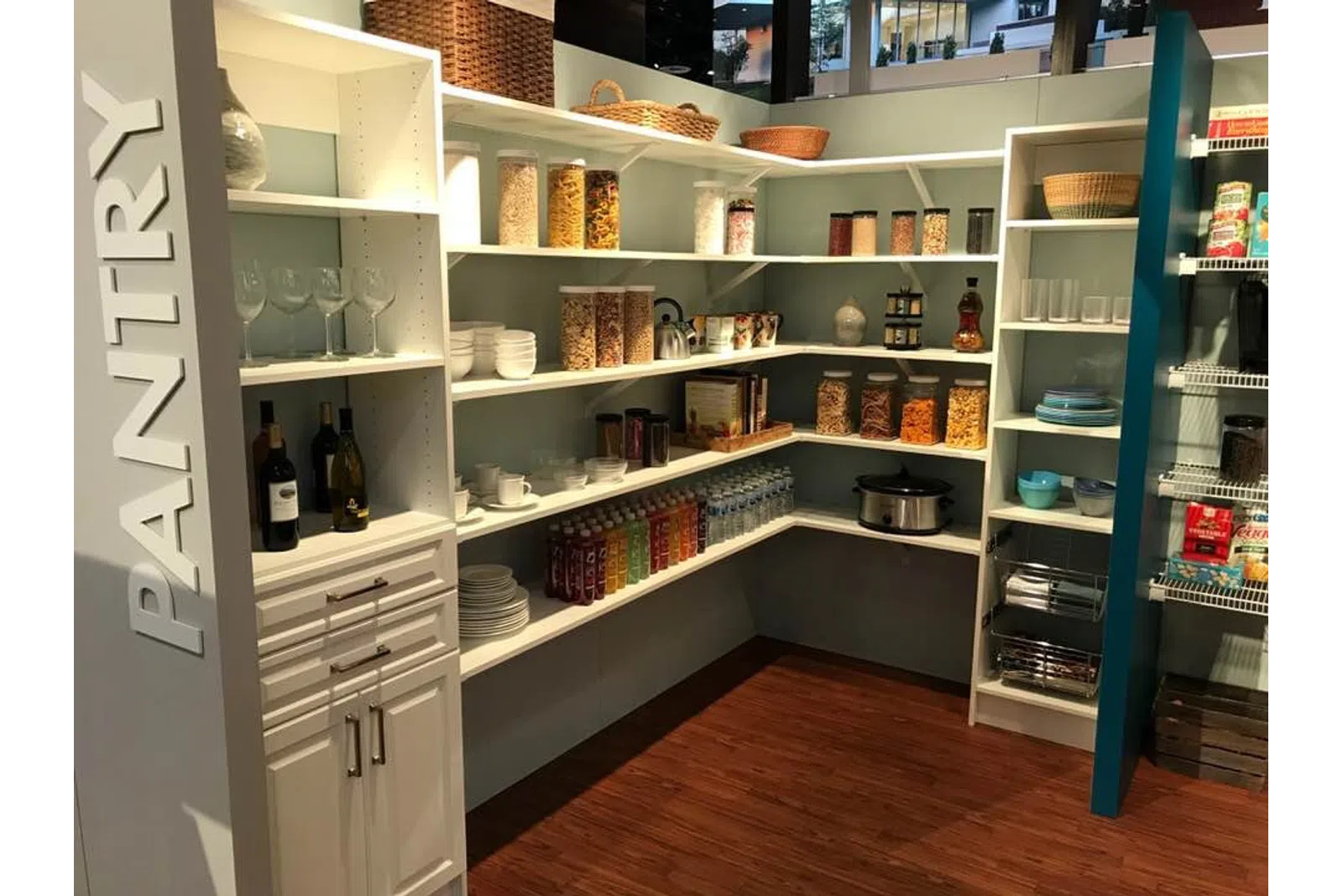 Organized pantry with white shelves holding jars, bottles, dishes, and baskets on wood flooring under bright lighting