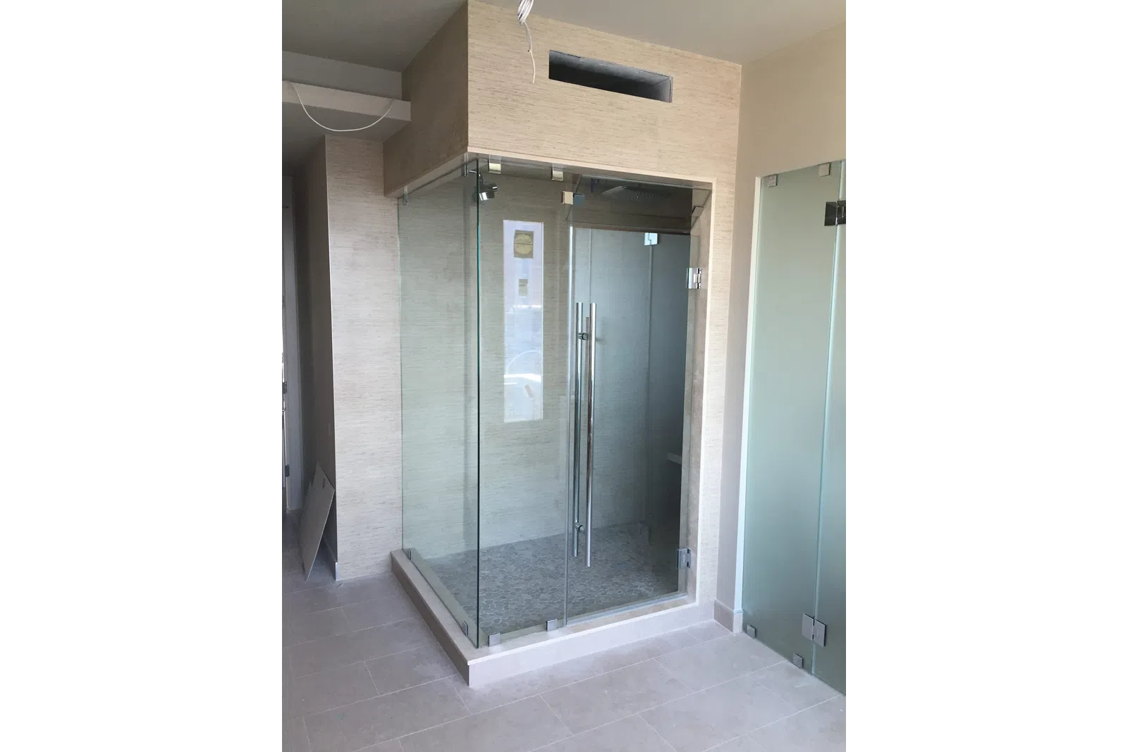 Modern walk-in shower with glass doors in a beige tiled bathroom under construction.