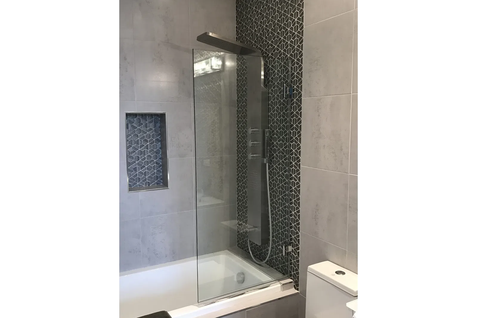Modern bathroom shower area with glass partition, geometric tile accent, and sleek stainless steel shower panel.