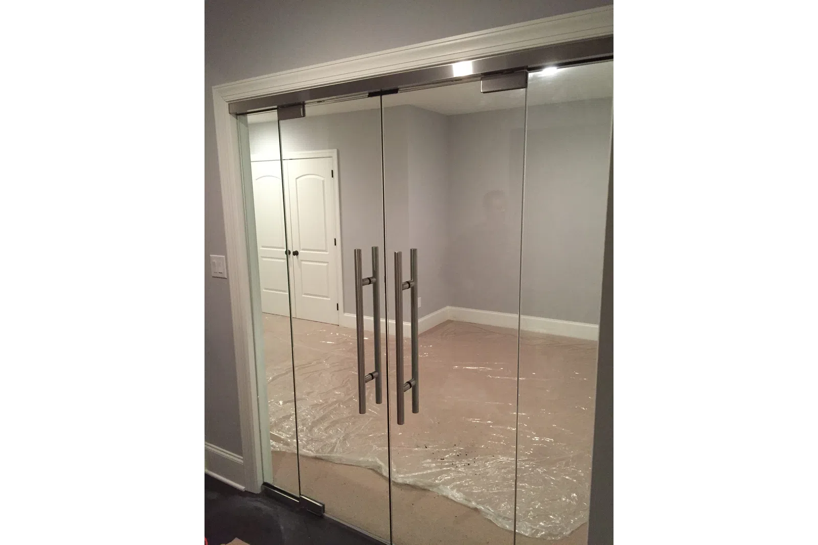 Modern glass double doors with metal handles leading into a room with plastic-covered floor and closed white doors.