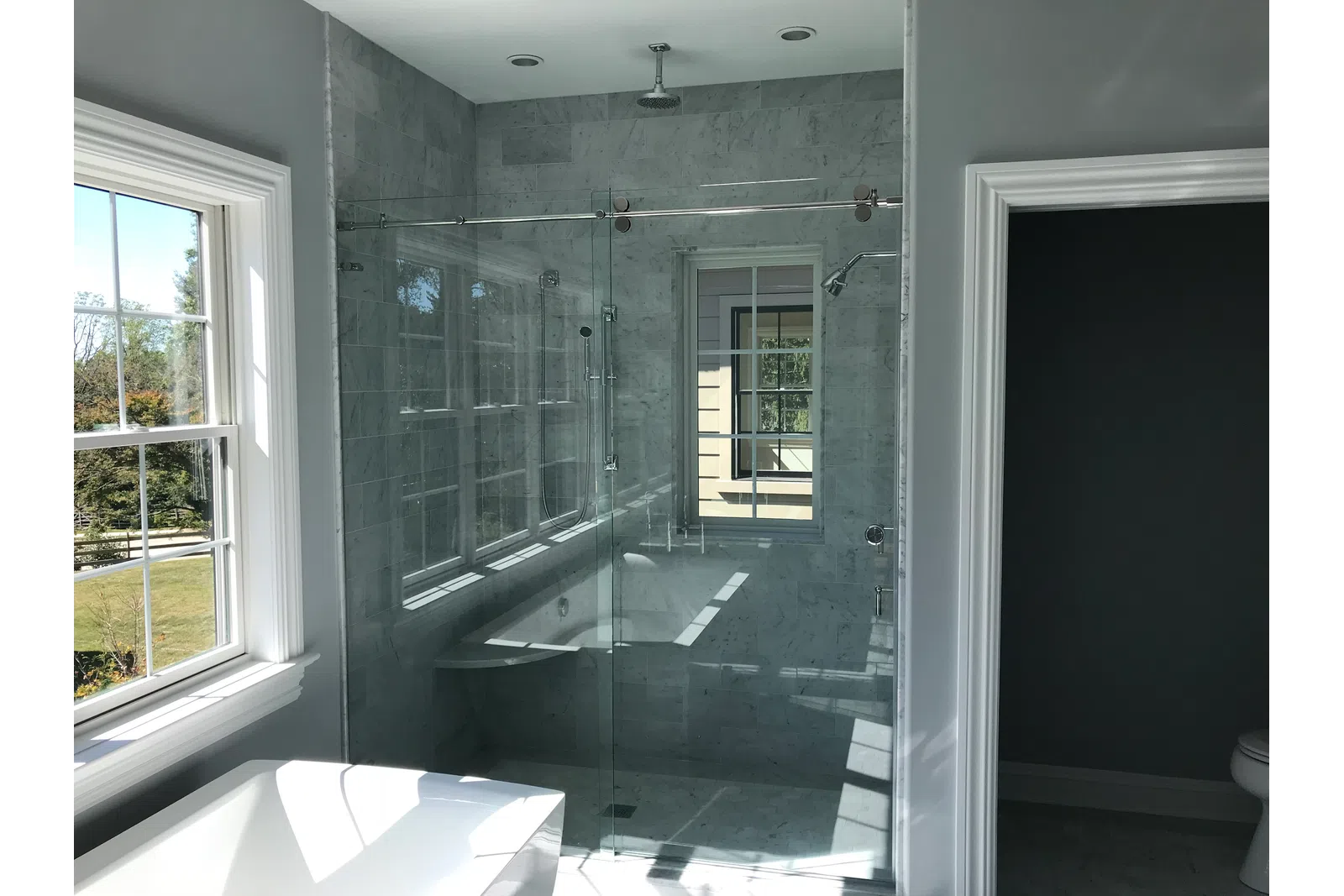 Modern bathroom with glass-enclosed shower, marble tiles, bathtub, and large windows with natural light.