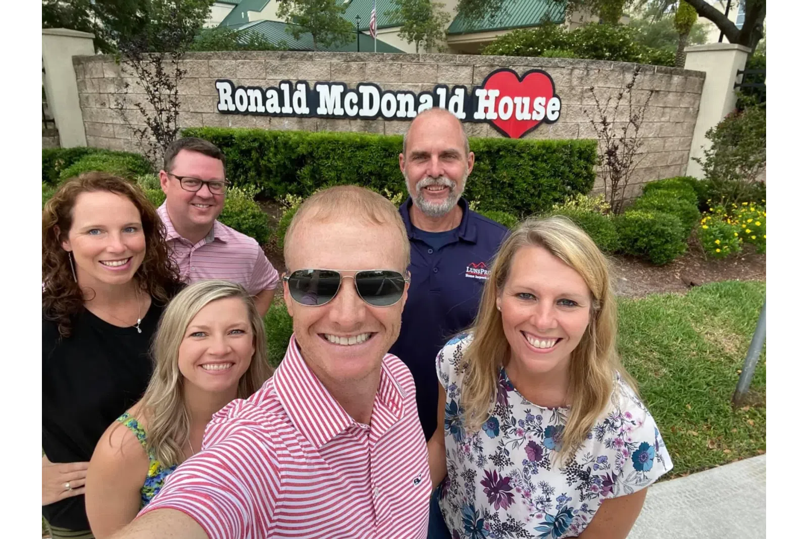 Group selfie of six smiling adults in front of Ronald McDonald House sign outdoors on a sunny day