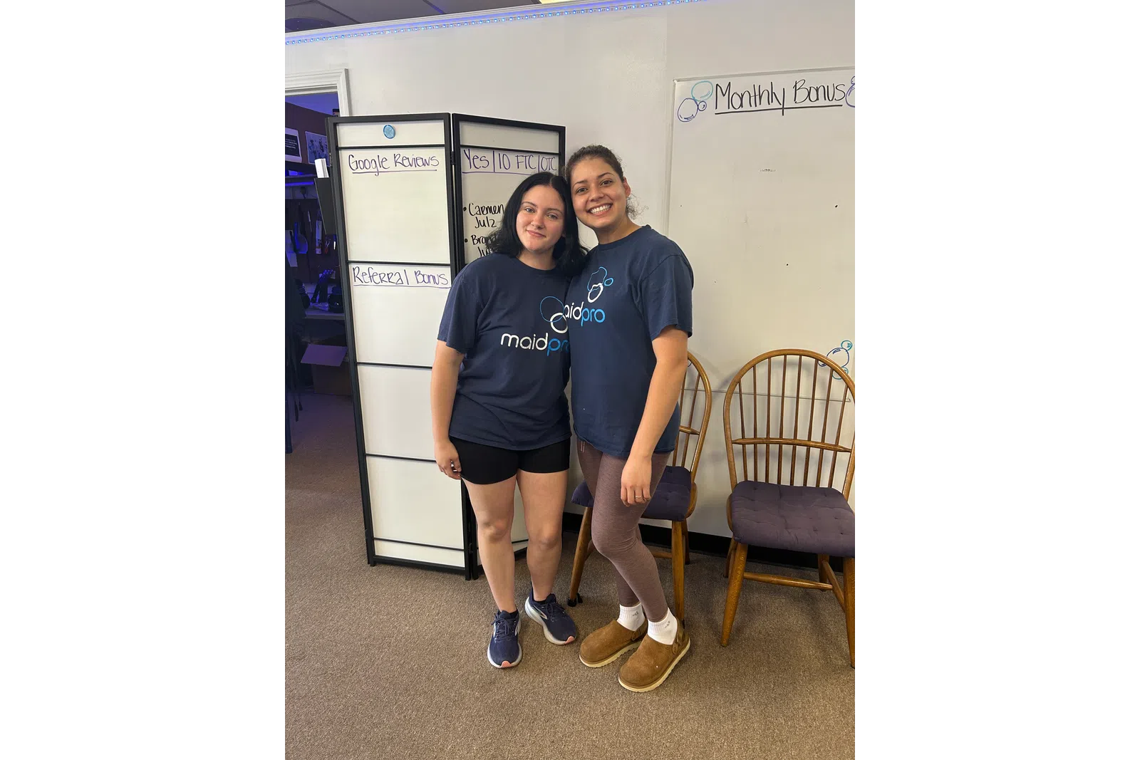 Two women smiling wearing MaidPro t-shirts standing in front of office whiteboards and chairs.