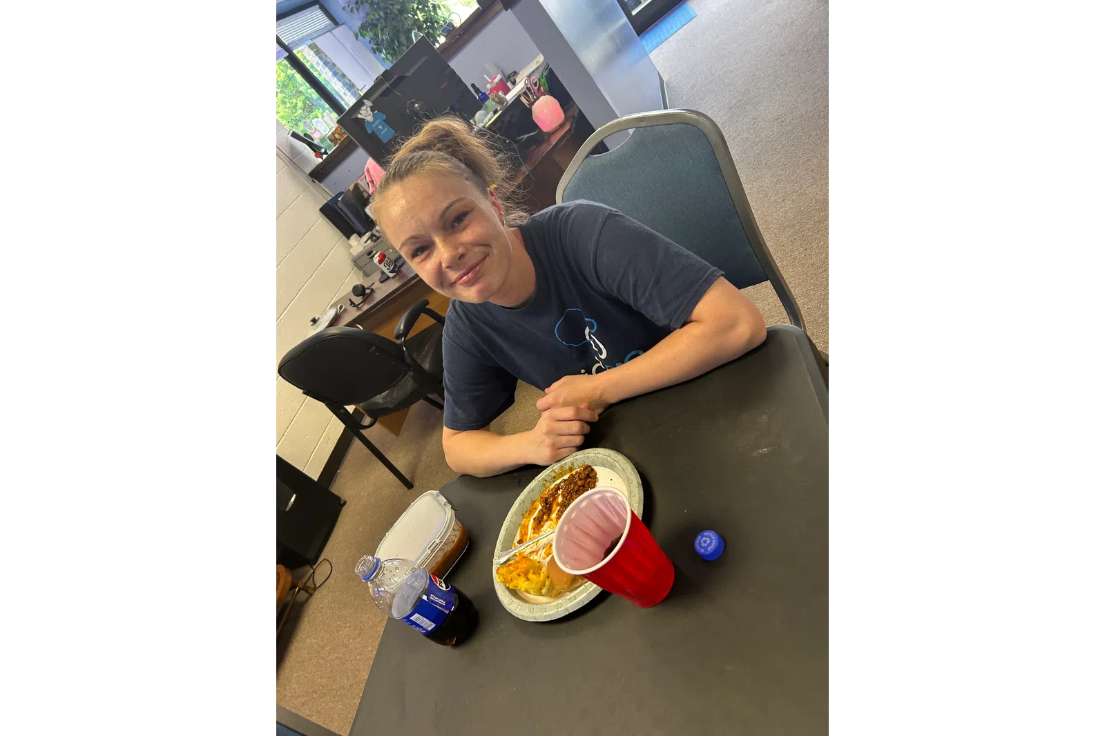 Young woman smiling at a table with a plate of food, a red cup, and a soda bottle in an office setting.