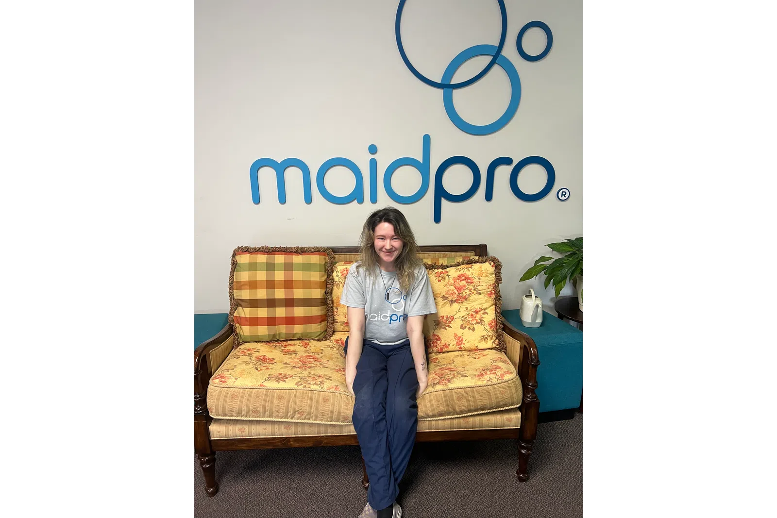 Smiling woman in MaidPro shirt sitting on floral couch under MaidPro logo with decorative pillows and plant nearby