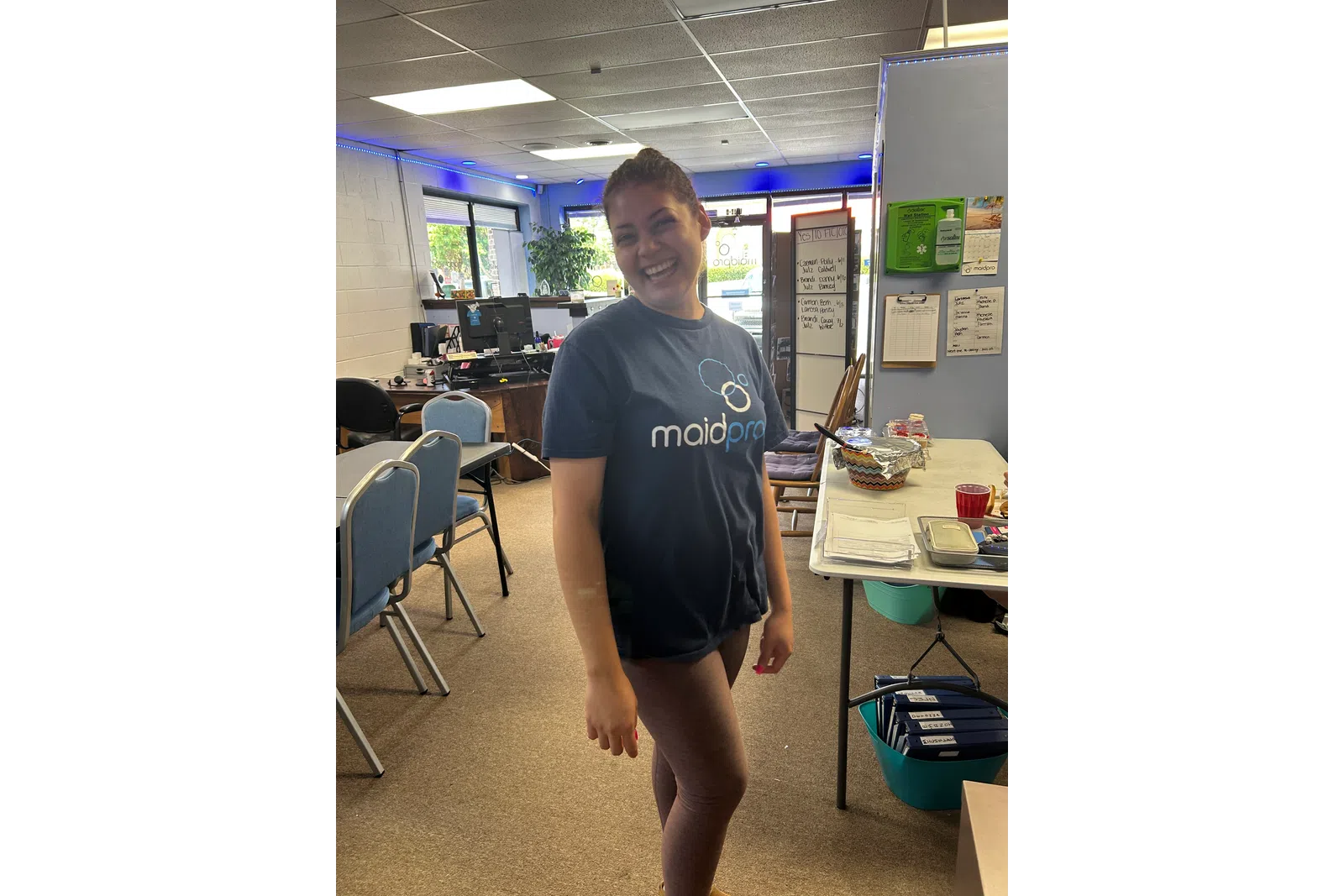 Smiling woman wearing a MaidPro shirt stands in a bright office with chairs, a table, and documents.