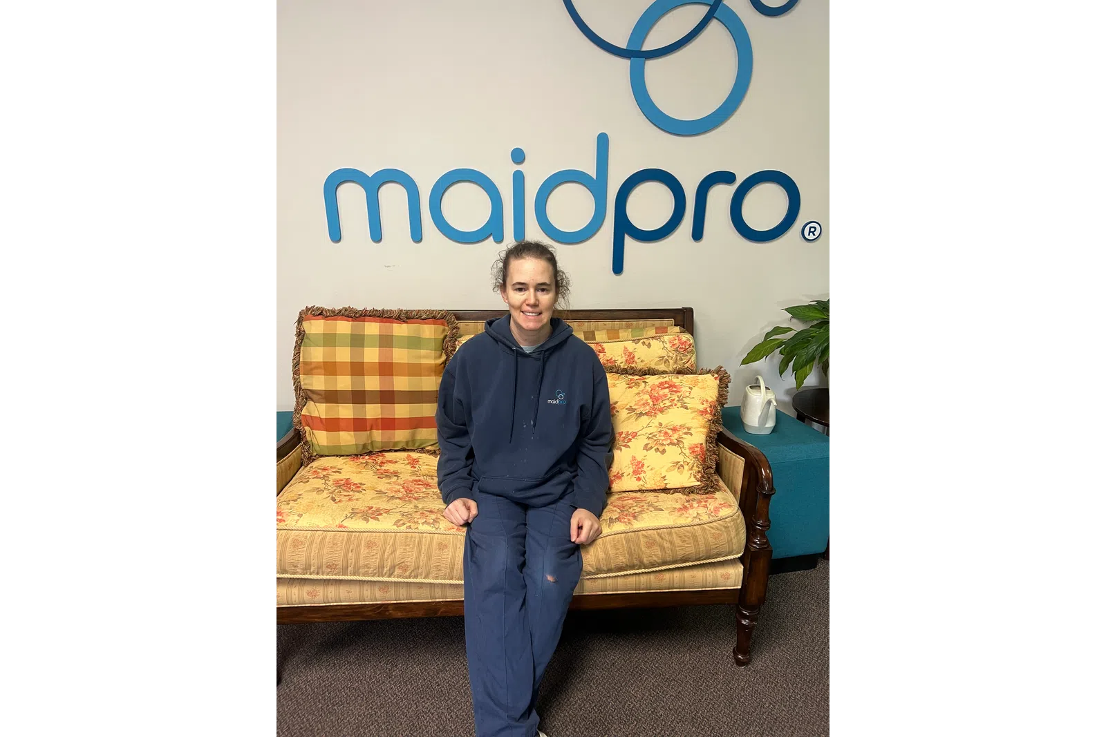 Woman in a MaidPro uniform sitting on a vintage floral sofa under a MaidPro logo on a white wall.