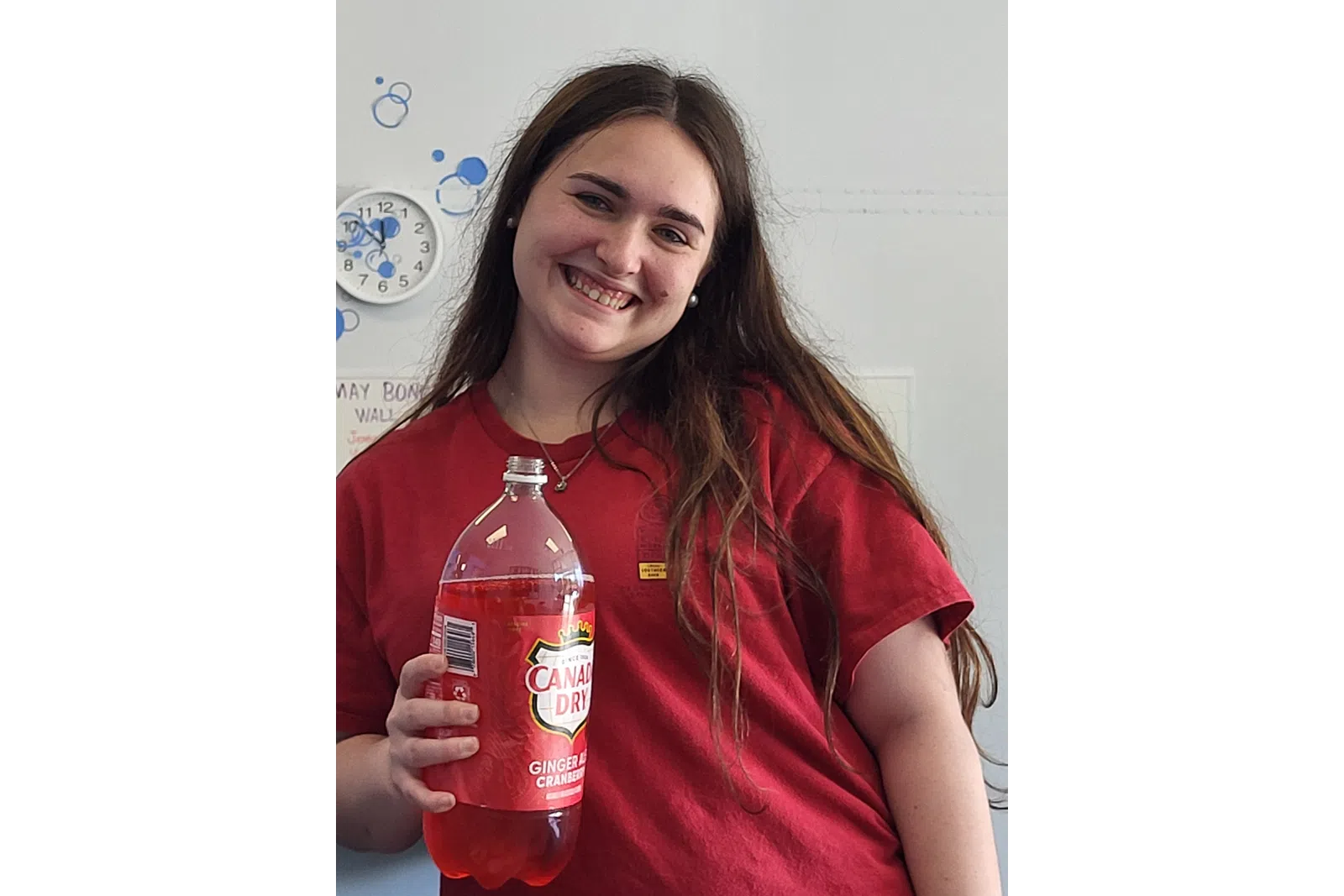 Smiling young woman in red shirt holding a Canada Dry Ginger Ale Cranberry soda bottle indoors.