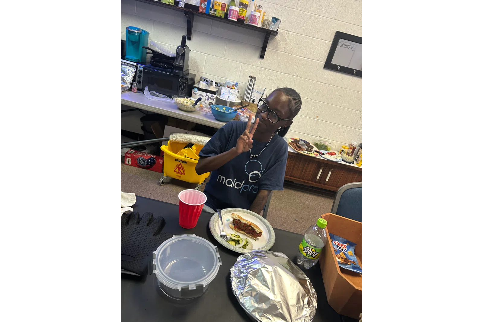 Person wearing MaidPro shirt making peace sign while sitting at table with food and drink in break room.