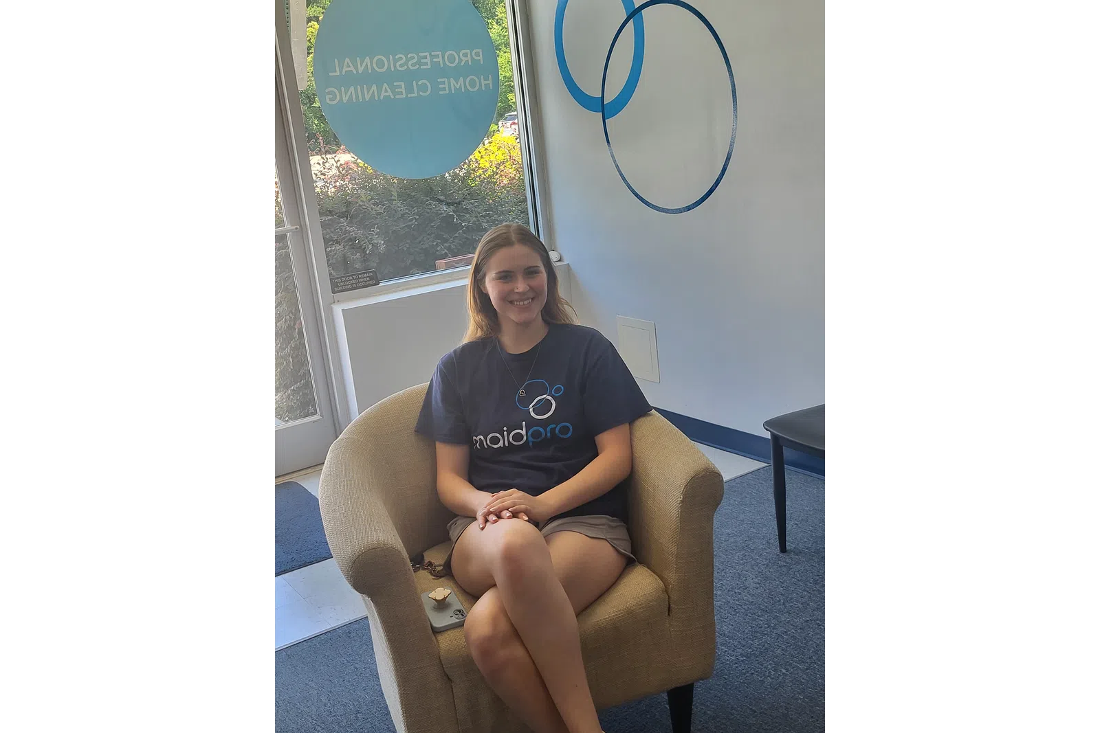 Smiling woman wearing MaidPro shirt sitting comfortably in an armchair in a professional home cleaning office.