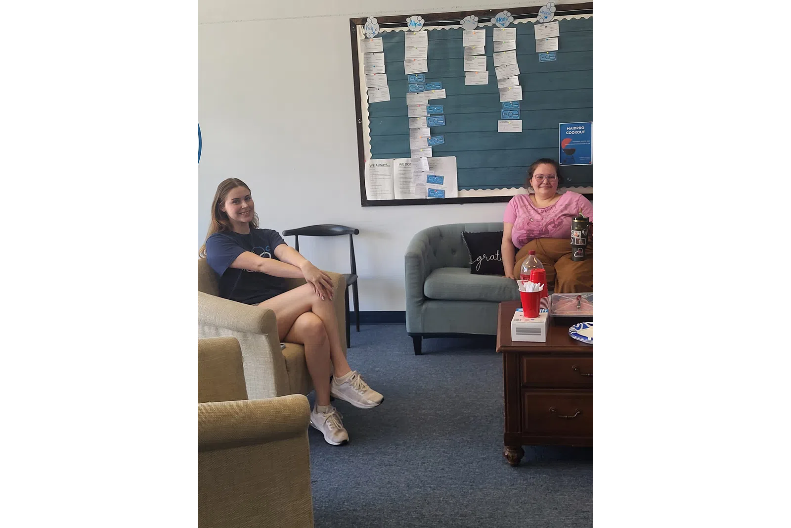 Two women sitting in a casual office lounge with a bulletin board and coffee table with snacks.
