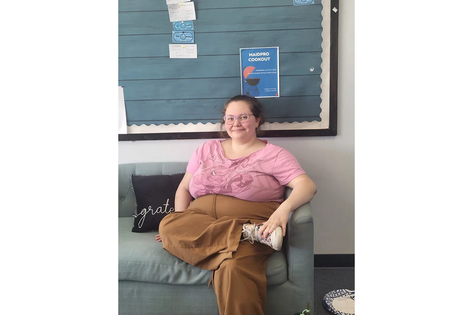 Smiling woman in pink shirt and brown pants sitting cross-legged on light gray couch with bulletin board background.