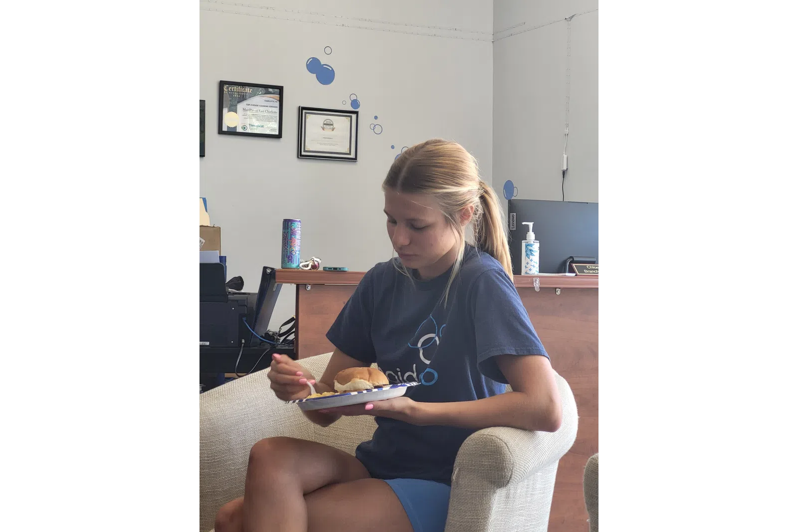 Young woman in navy shirt sitting on beige chair eating from a plate with sandwich and food indoors.