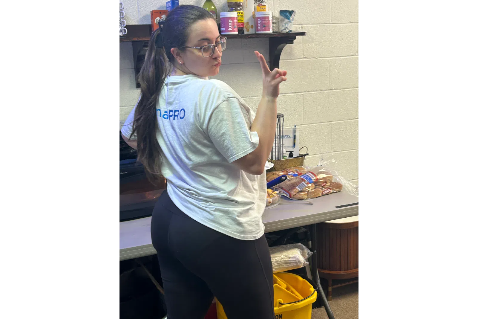 Woman in glasses and white T-shirt posing in a kitchen area with food items and cleaning supplies visible.