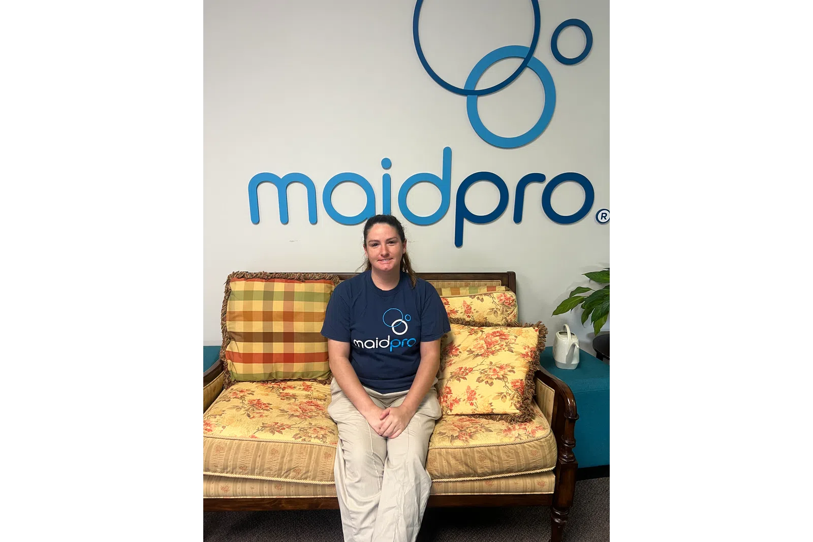 Woman in MaidPro shirt sitting on floral couch under MaidPro logo on white wall.