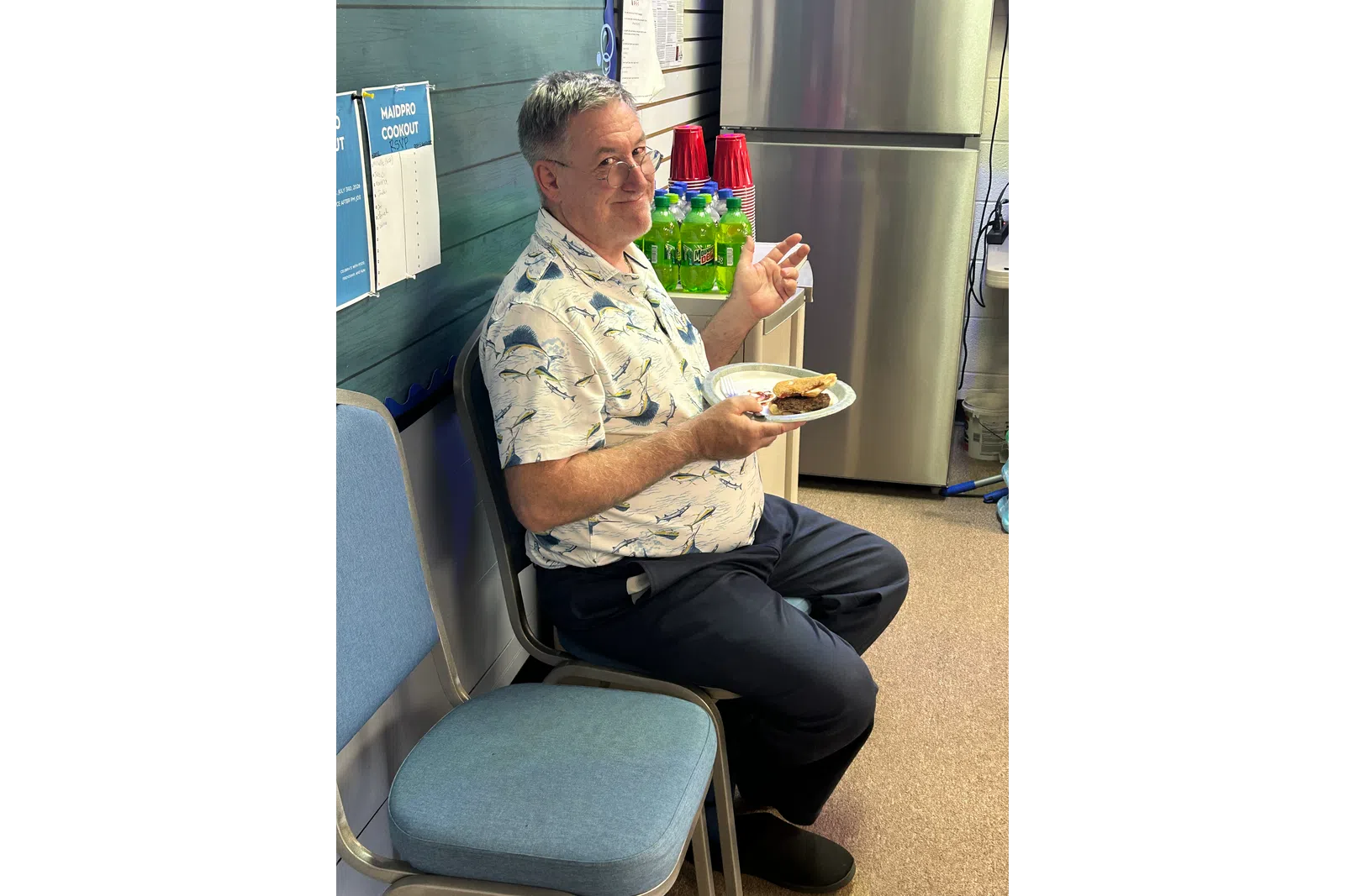 Man with glasses in patterned shirt sitting and eating a burger at an indoor gathering with drinks and red cups nearby.