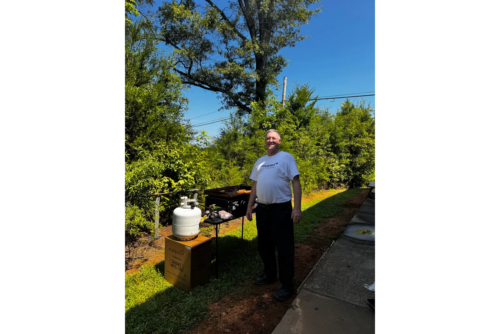 Man standing near a grill with propane tank in a sunny backyard surrounded by green trees and blue sky.