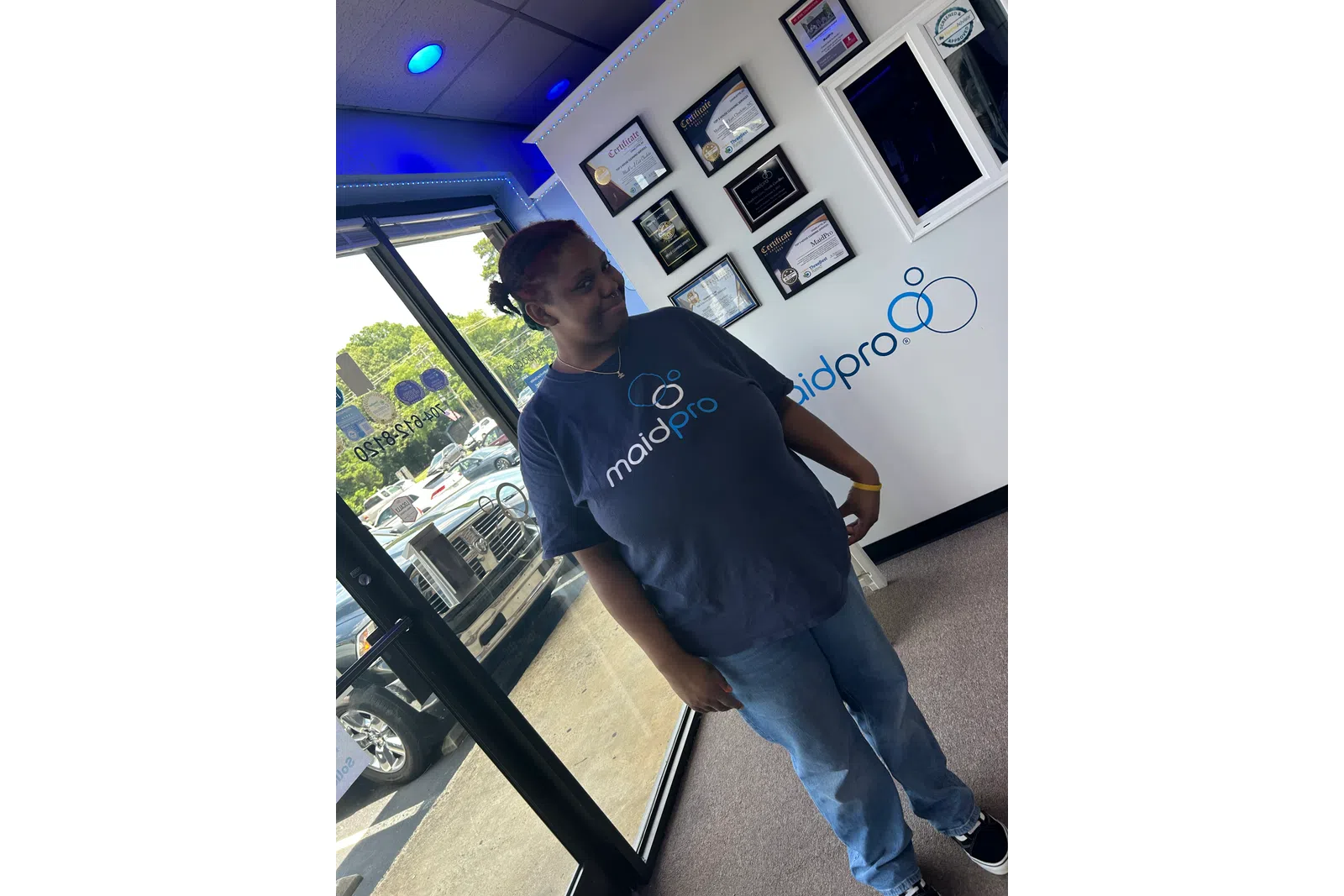 Woman standing inside MaidPro office wearing a MaidPro t-shirt near certificates and window.