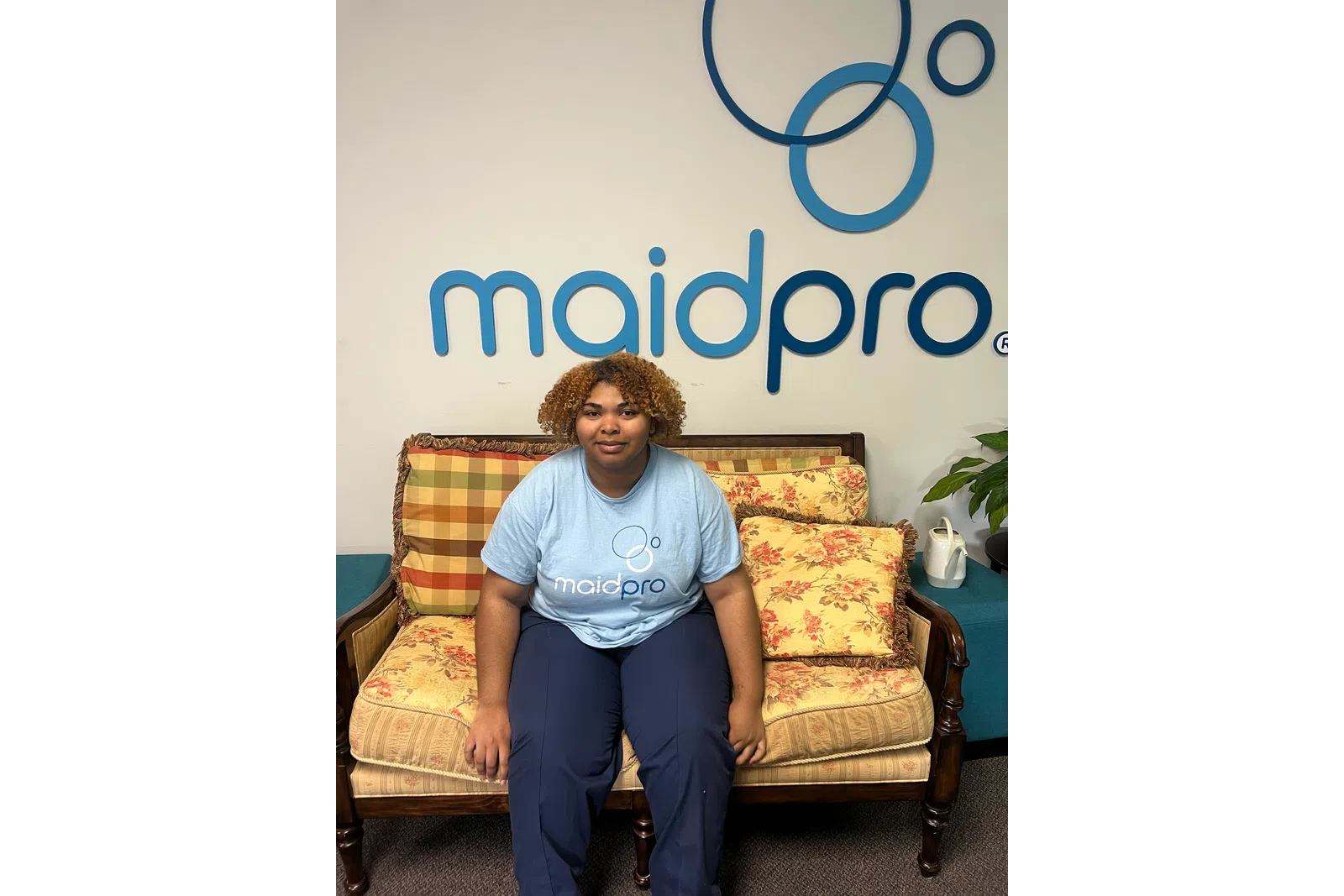 Woman with curly hair sitting on a floral patterned sofa in front of a MaidPro logo wall sign.