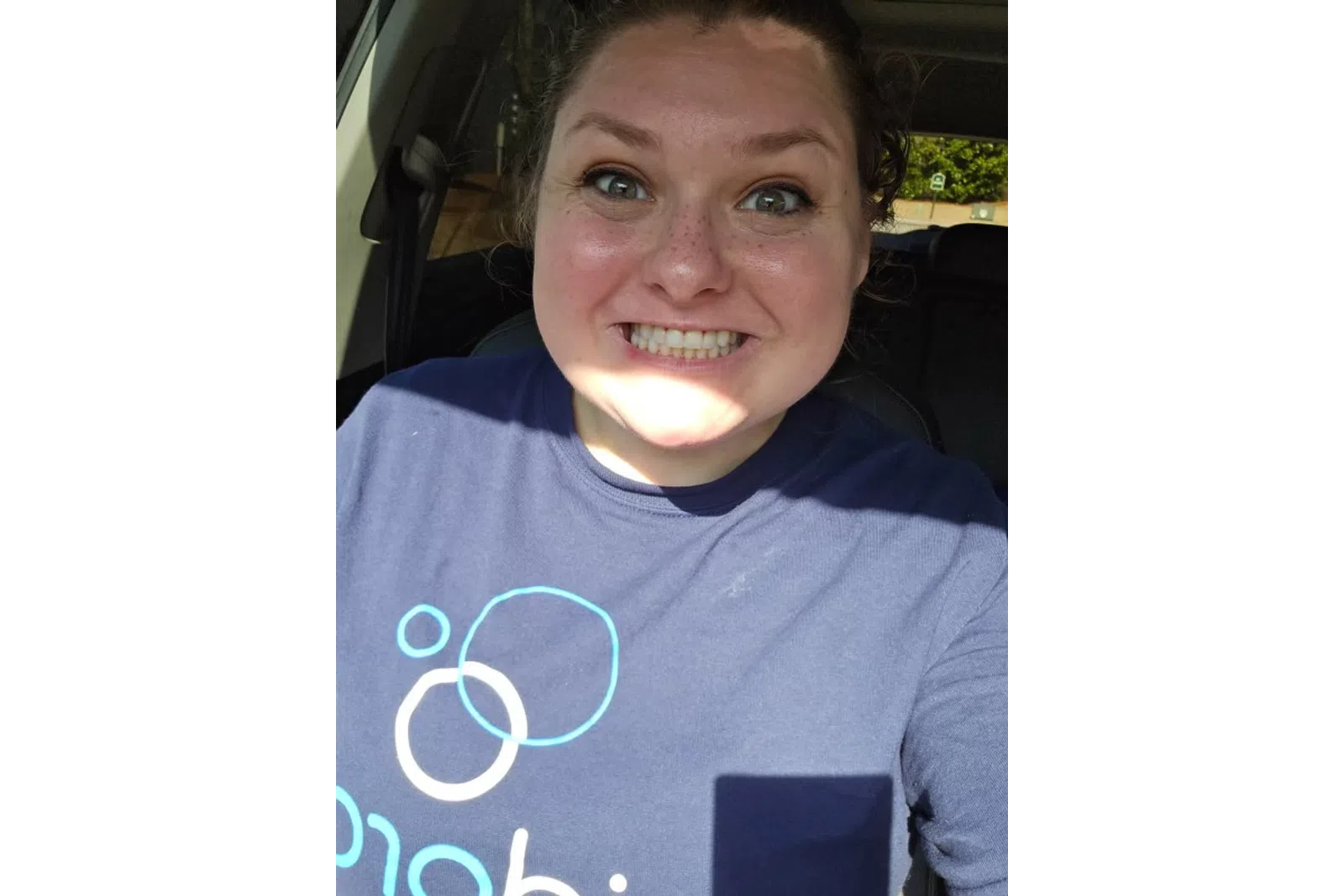 Smiling woman with freckles wearing a blue shirt with circular patterns sitting inside a car.