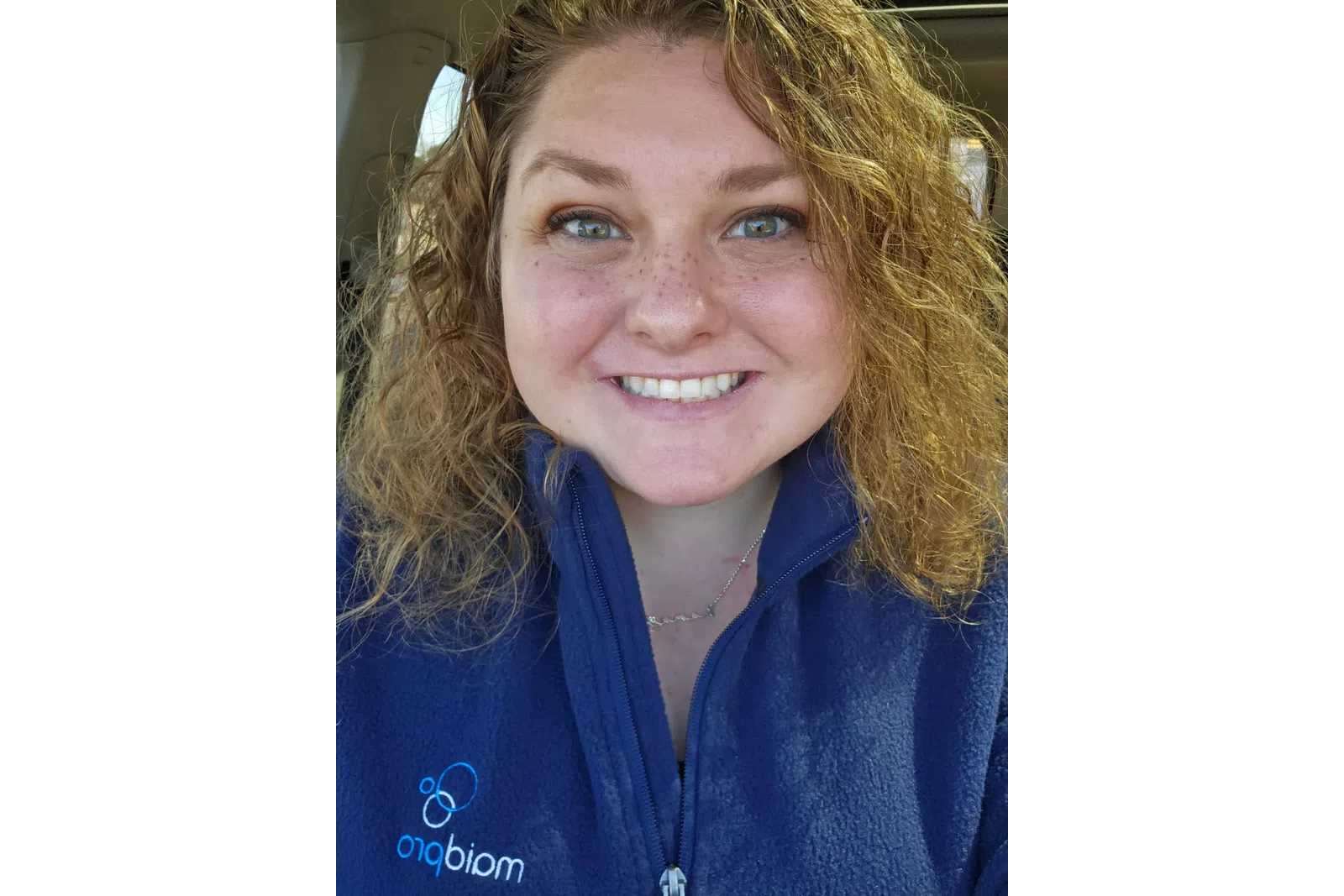 Smiling woman with curly hair wearing a blue zip-up jacket with a logo, sitting inside a car.