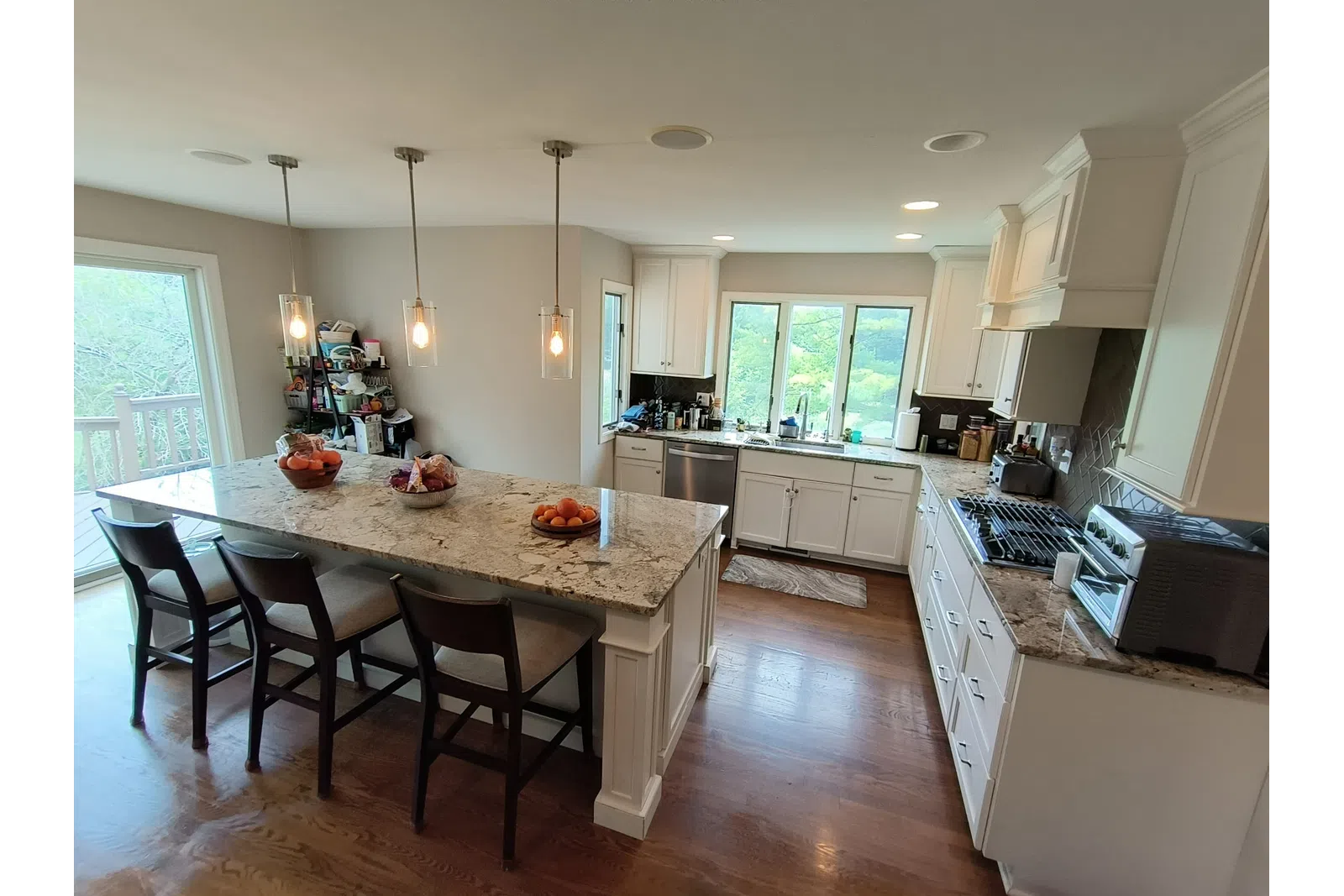 Modern kitchen with white cabinets, granite island countertop, wooden floor, and large windows overlooking greenery.