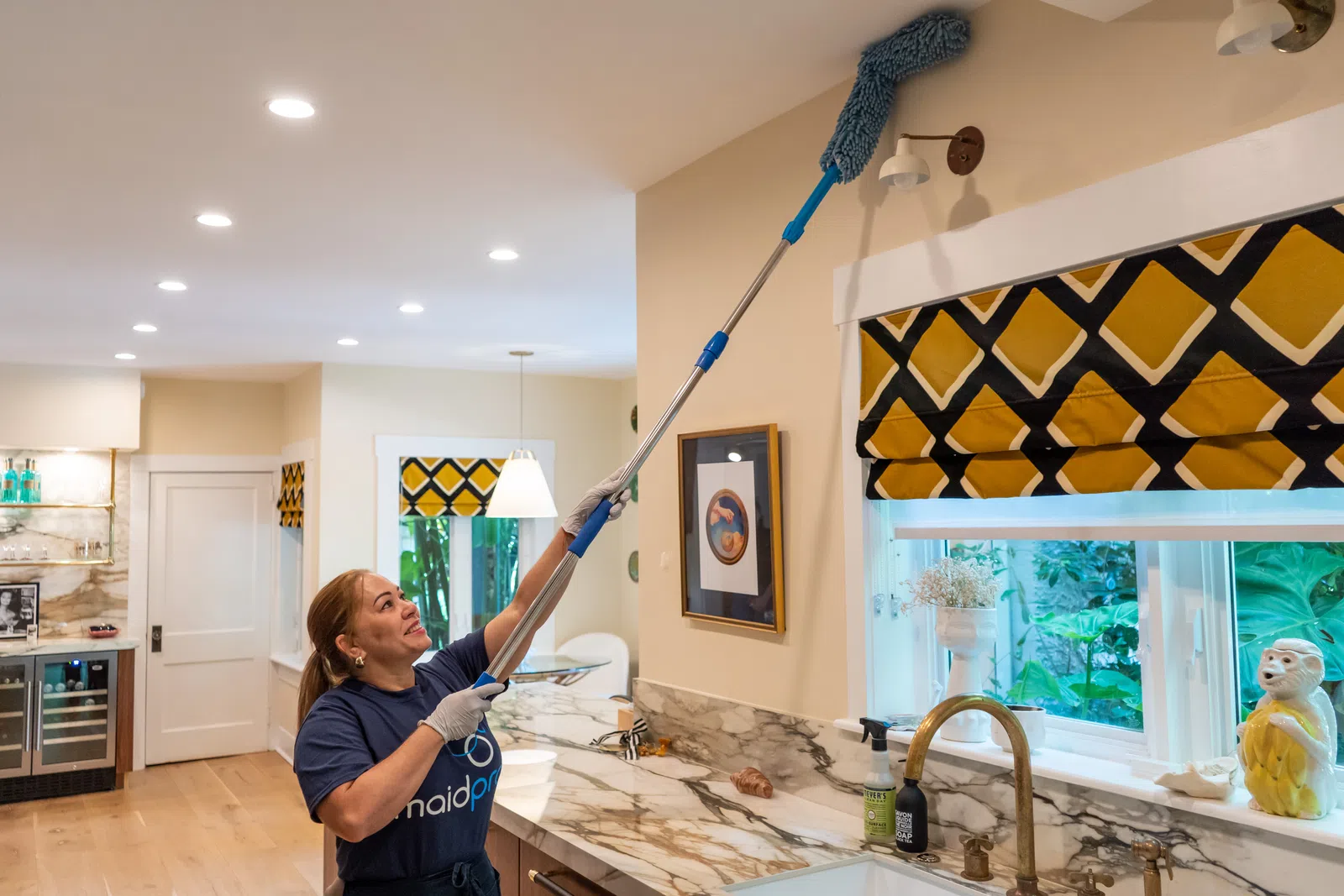 Woman using an extendable duster to clean ceiling corners in a modern kitchen with marble countertops and gold fixtures