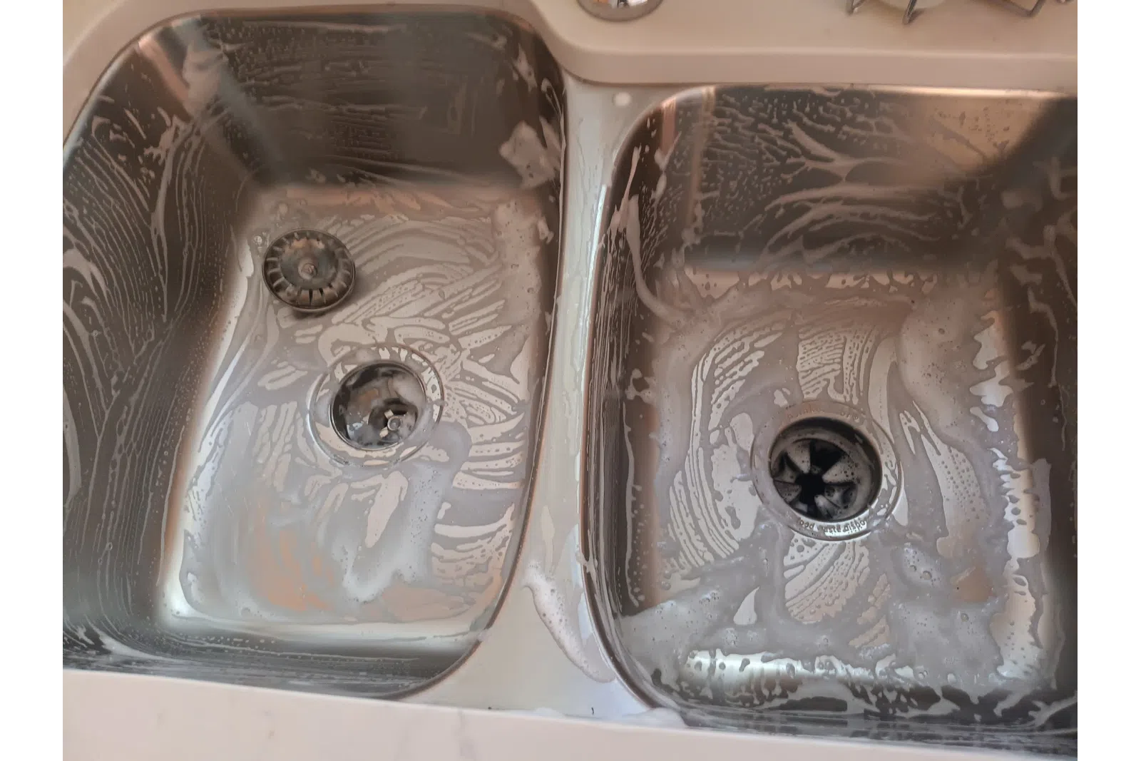 Two stainless steel kitchen sinks with soap suds and cleaning residue inside them.