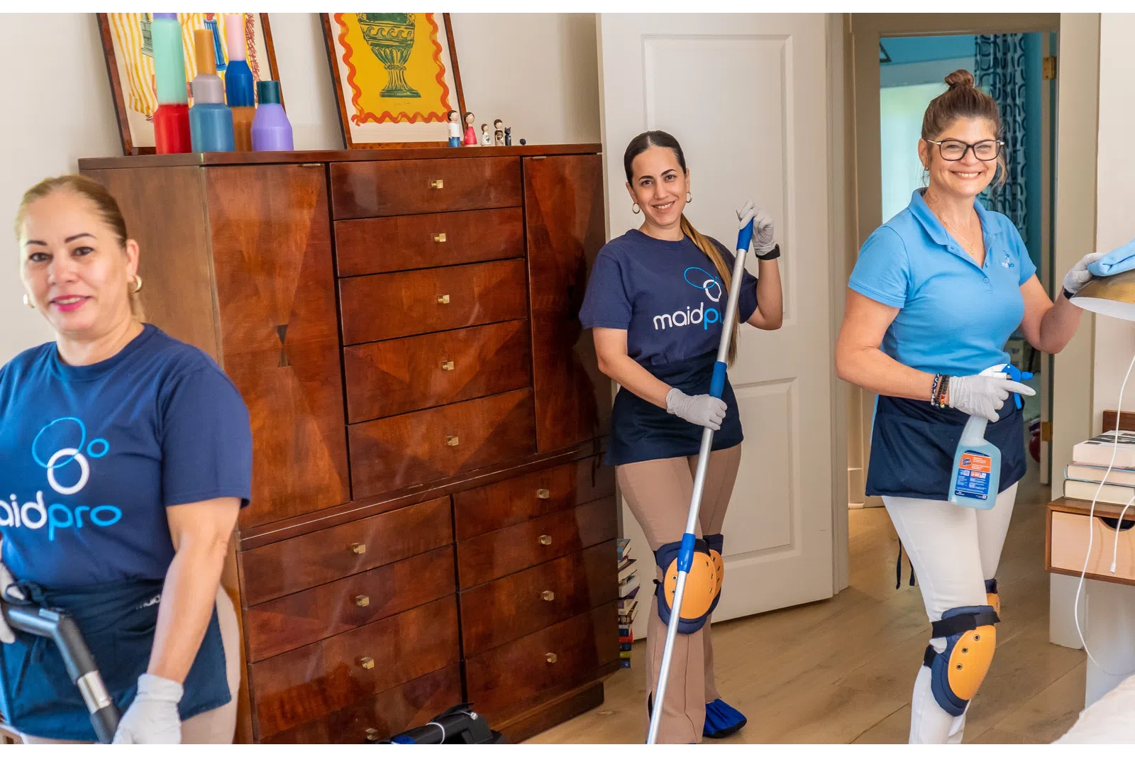 Three professional house cleaners wearing MaidPro uniforms cleaning a living room and smiling at the camera.