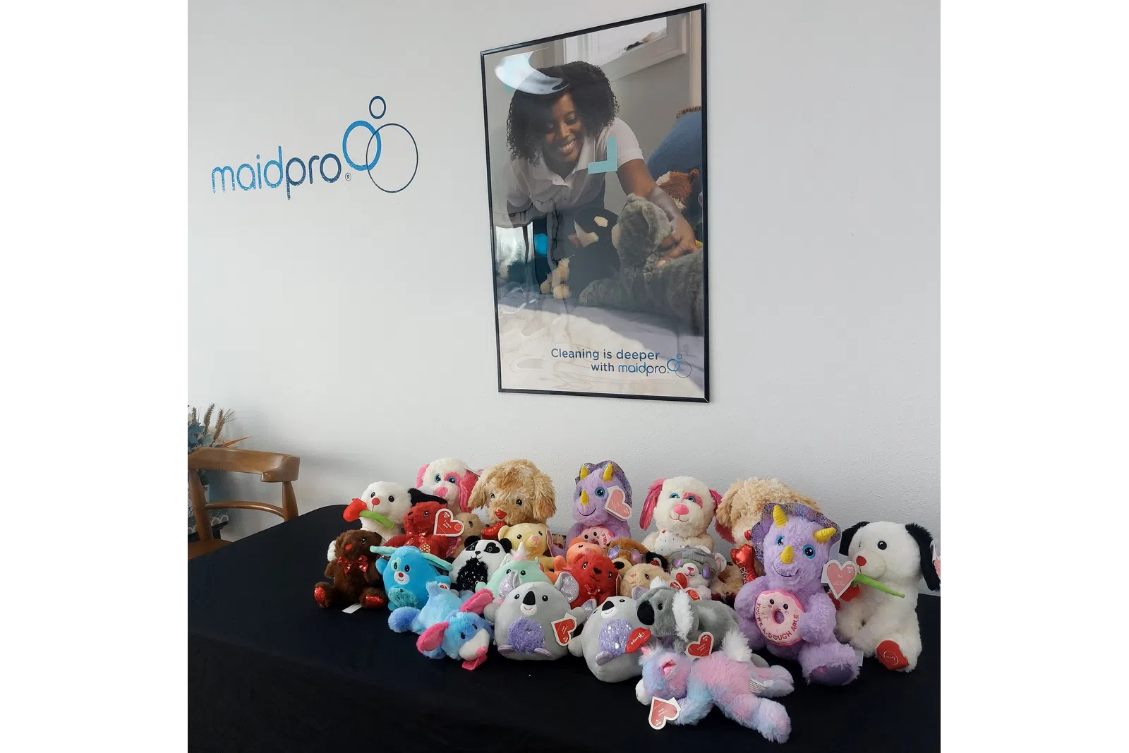 Variety of colorful stuffed animals displayed on a table under a MaidPro cleaning services poster and logo on the wall.