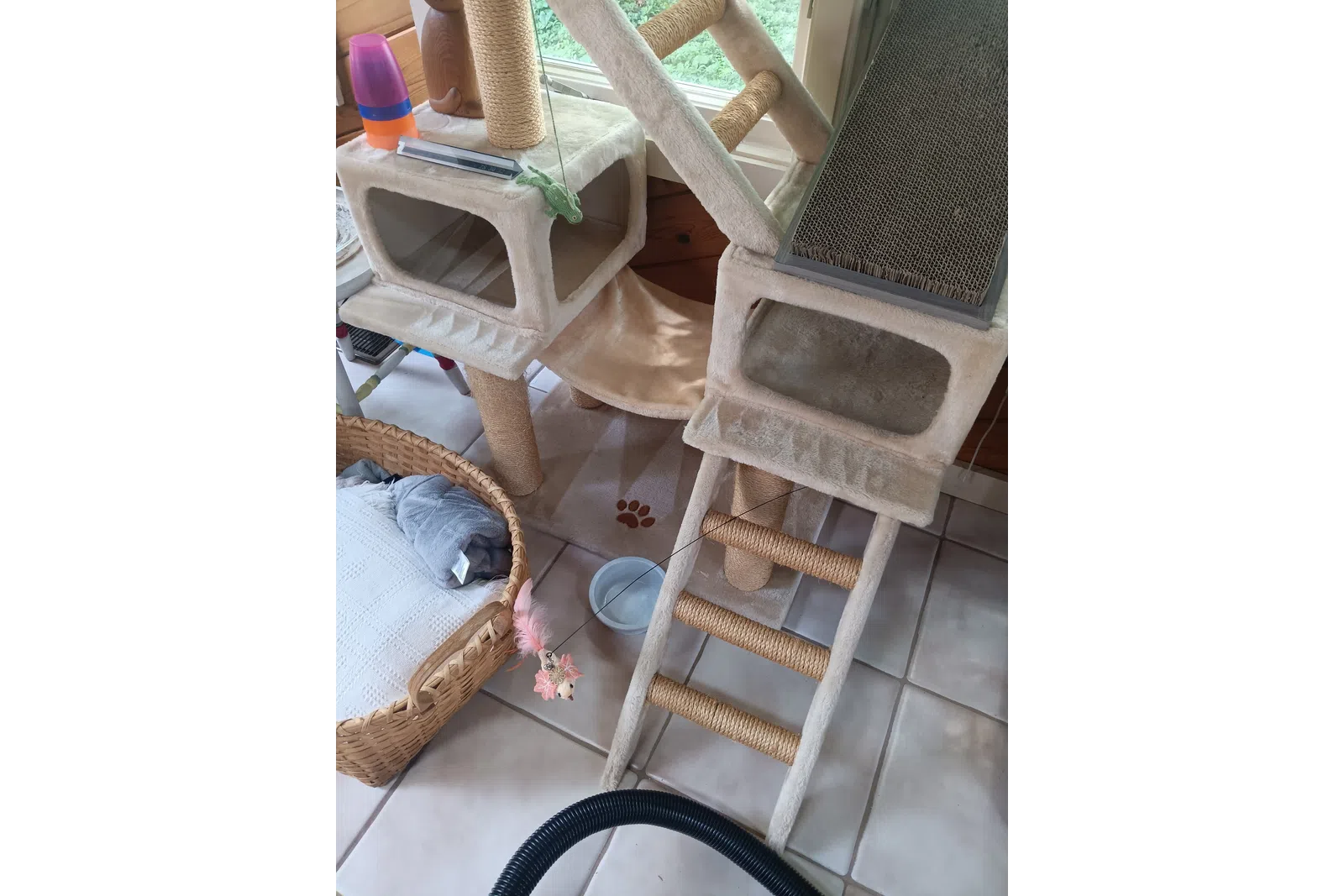 Beige cat tree with ladder, scratching pads, hanging toy, and cozy basket near a window on tiled floor.