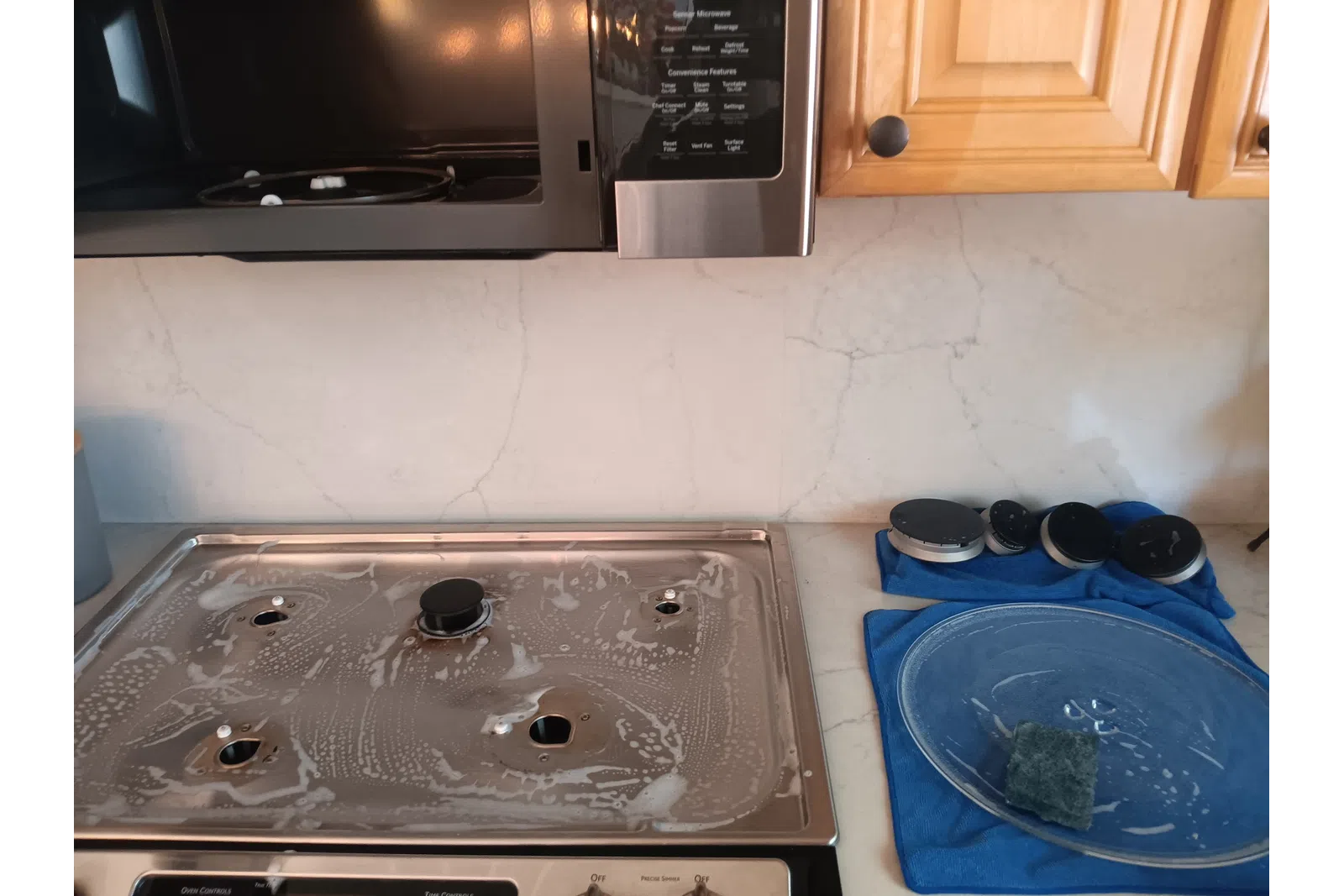 Cleaned gas stove top with soap suds, removed burner caps on blue towel, microwave and wooden cabinets in kitchen.