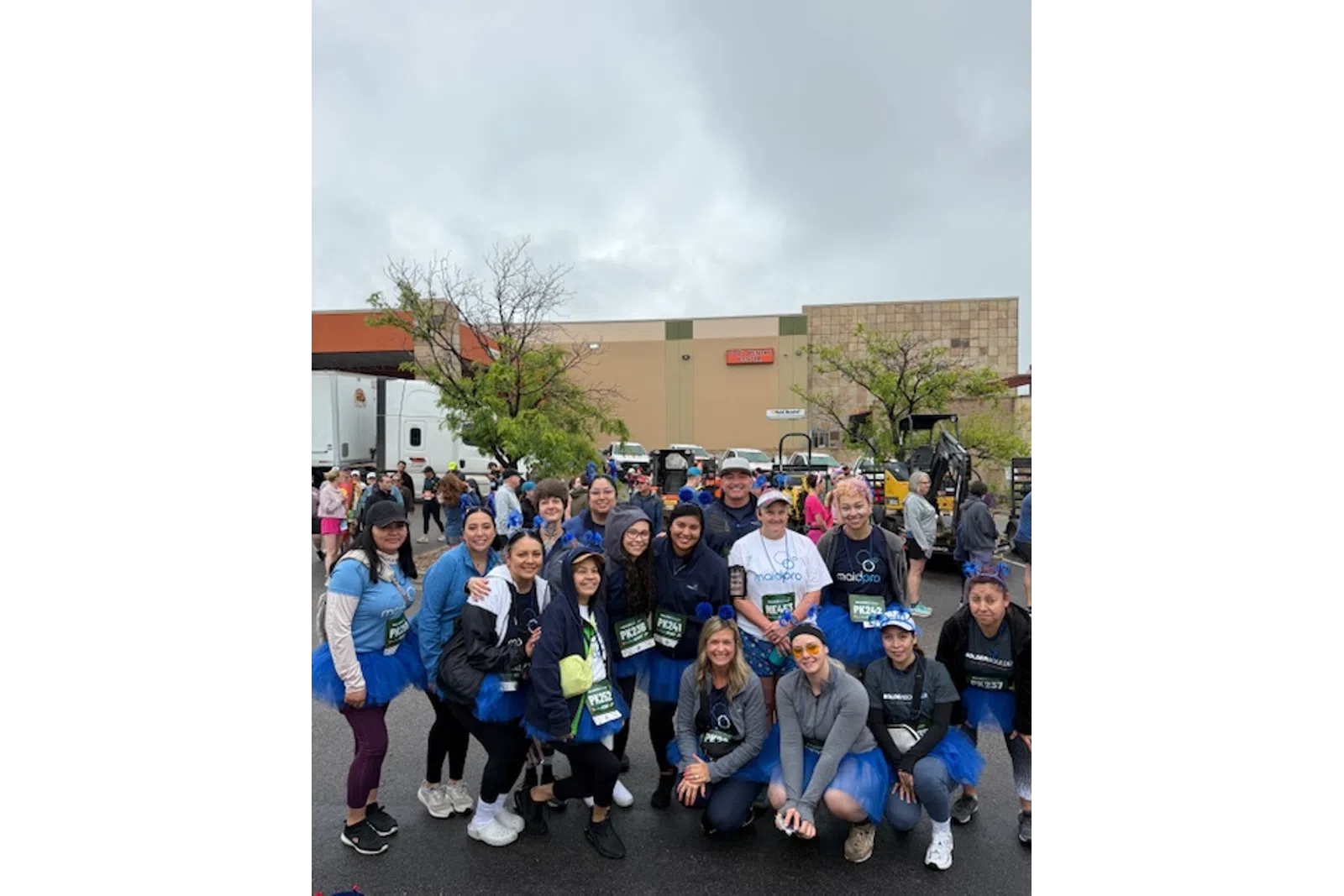 Group of smiling participants in blue tutus and race bibs posing outdoors before a community race event on a cloudy day.