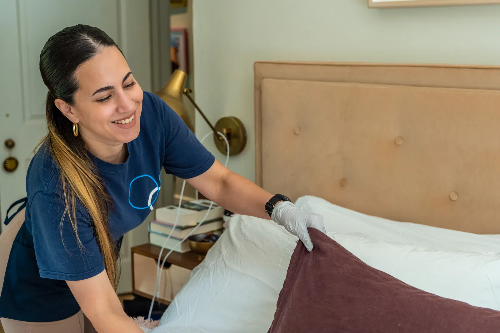 Smiling woman wearing gloves arranges pillows on a neatly made bed in a cozy bedroom setting.