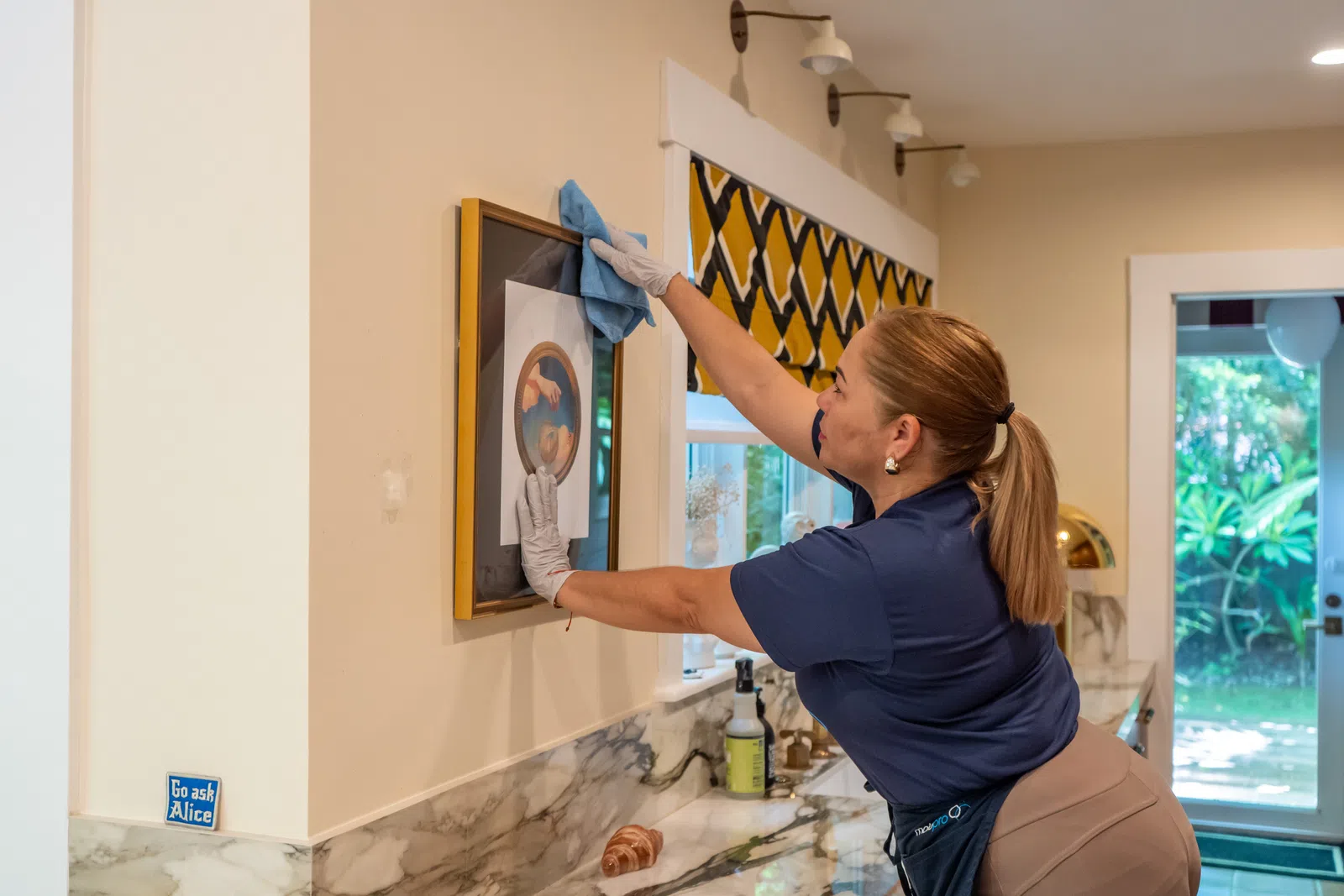 Woman wearing gloves cleans framed artwork on kitchen wall with blue cloth in bright room.
