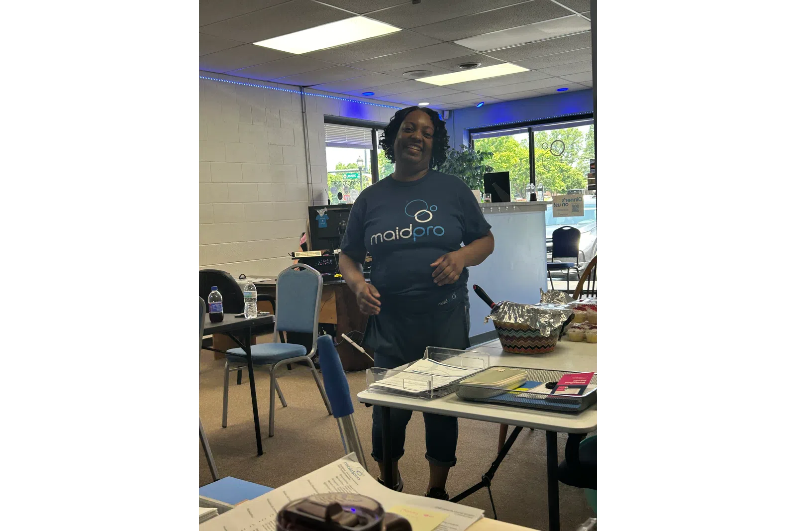 Smiling woman wearing a MaidPro t-shirt standing in a room with tables, chairs, and office supplies.