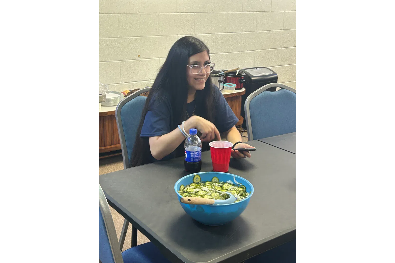 Young woman with long dark hair sitting at a table with a bowl of sliced cucumbers, red cup, and soda bottle.