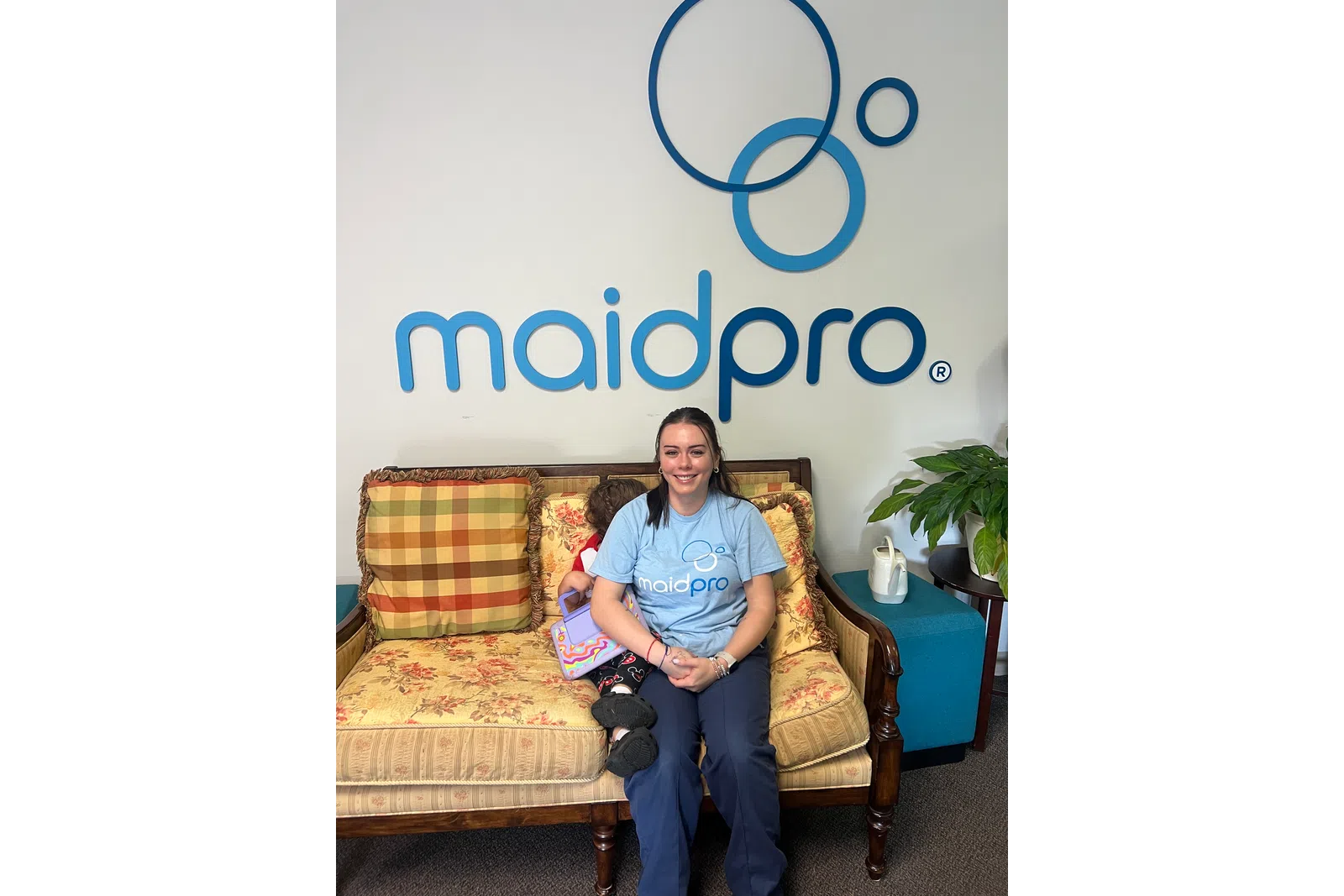 Smiling woman in maidpro shirt sitting on vintage sofa with child holding a toy, maidpro logo on wall behind.