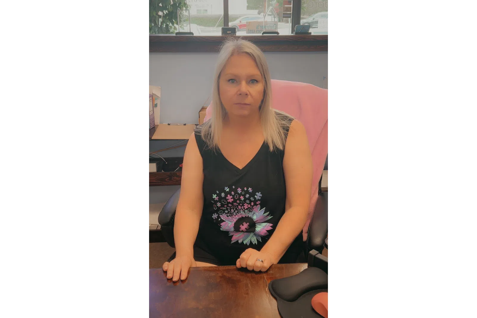 Woman with blonde hair wearing a black sleeveless shirt with colorful puzzle flower design sitting at a wooden desk.